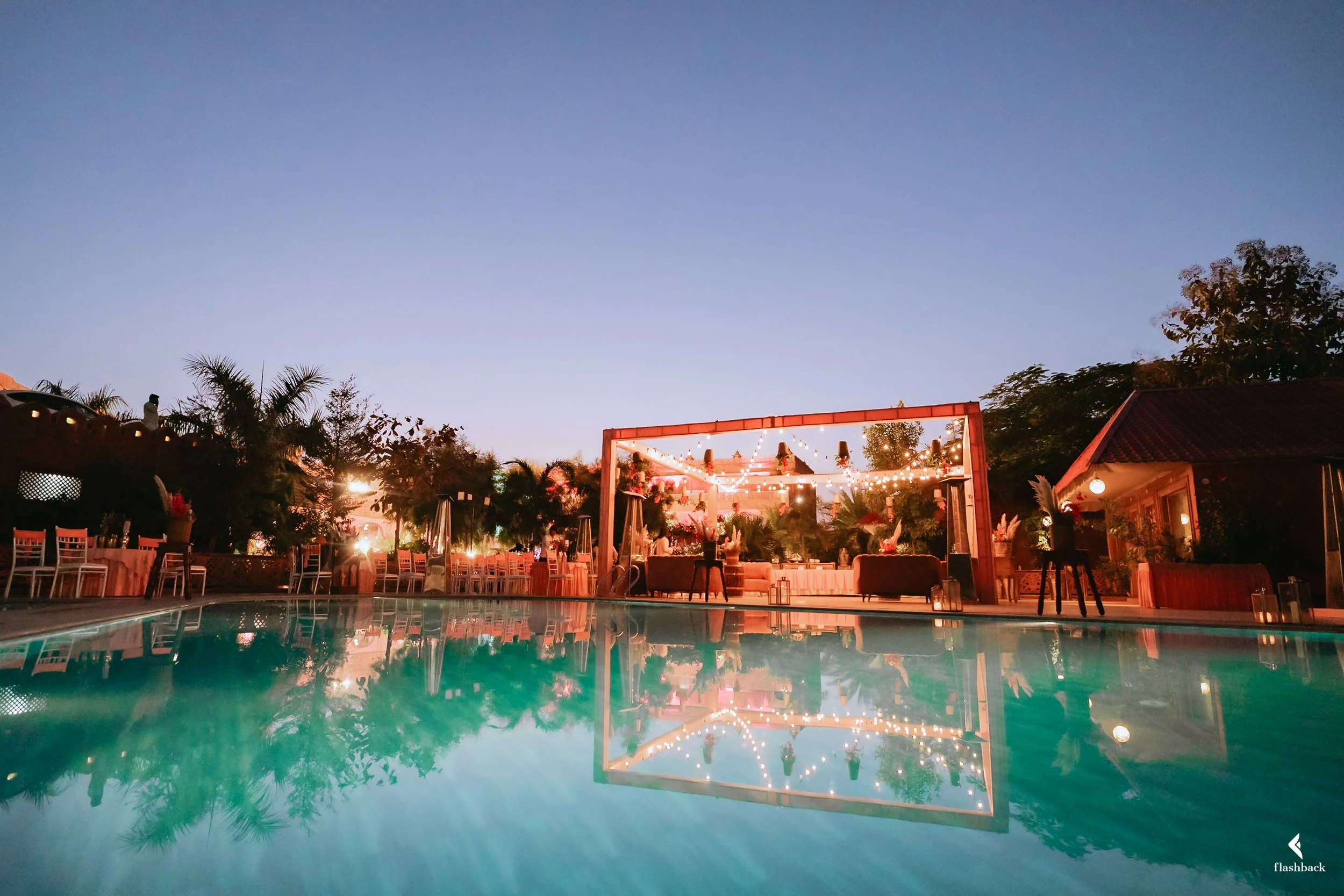 Outdoor party setup near a swimming pool at dusk with string lights, chairs, tables, palm trees, and a building with a red roof.