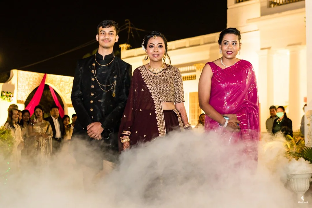 Three people at a nighttime celebration, dressed in traditional and formal attire, standing amidst a foggy or smoky setting outside a building with bright lighting.