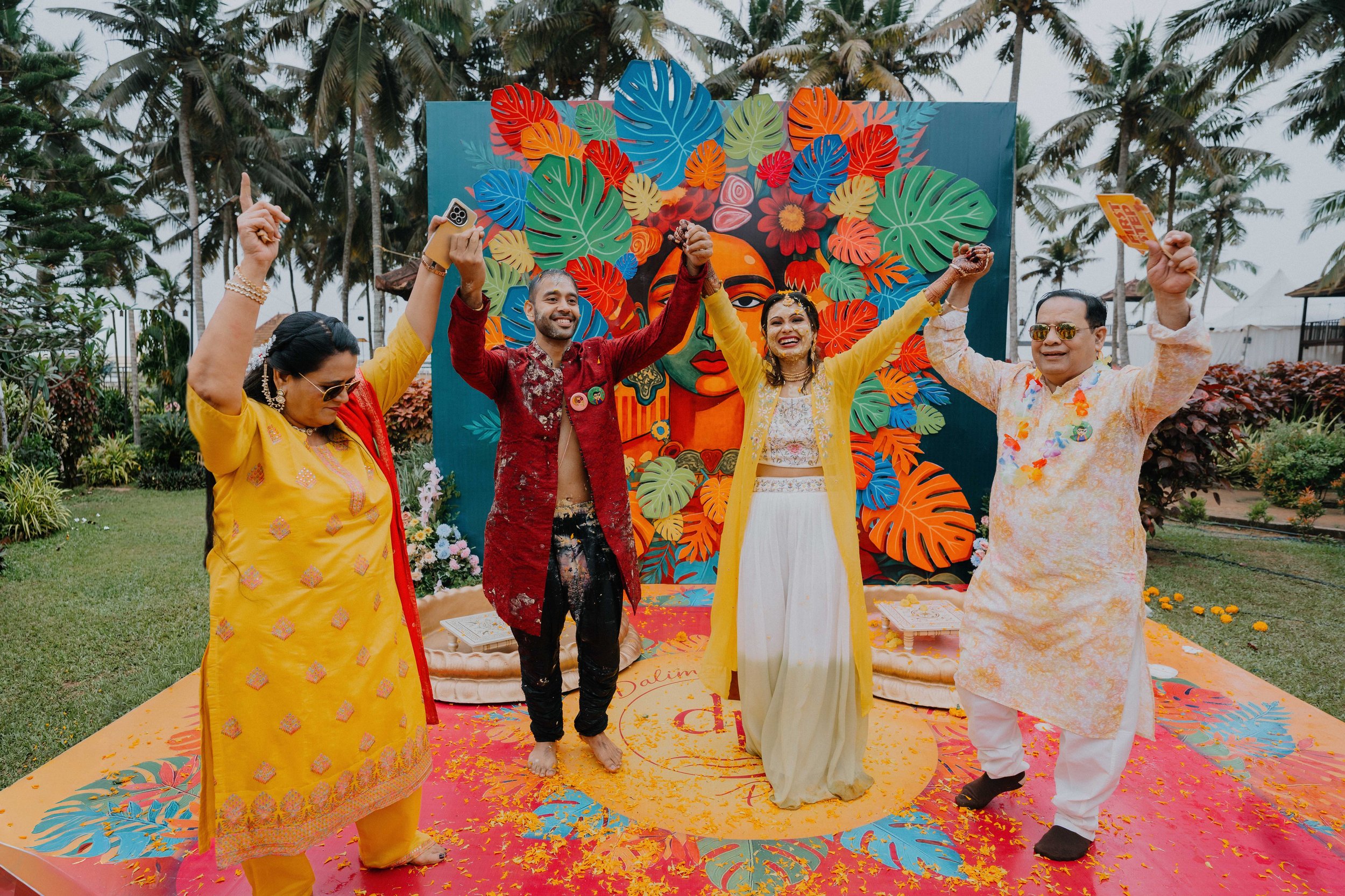 A group of five diverse people celebrating a wedding outdoors, with a colorful backdrop of tropical leaves and flowers, some are smiling, holding hands, and raising their arms, with petals on the ground.