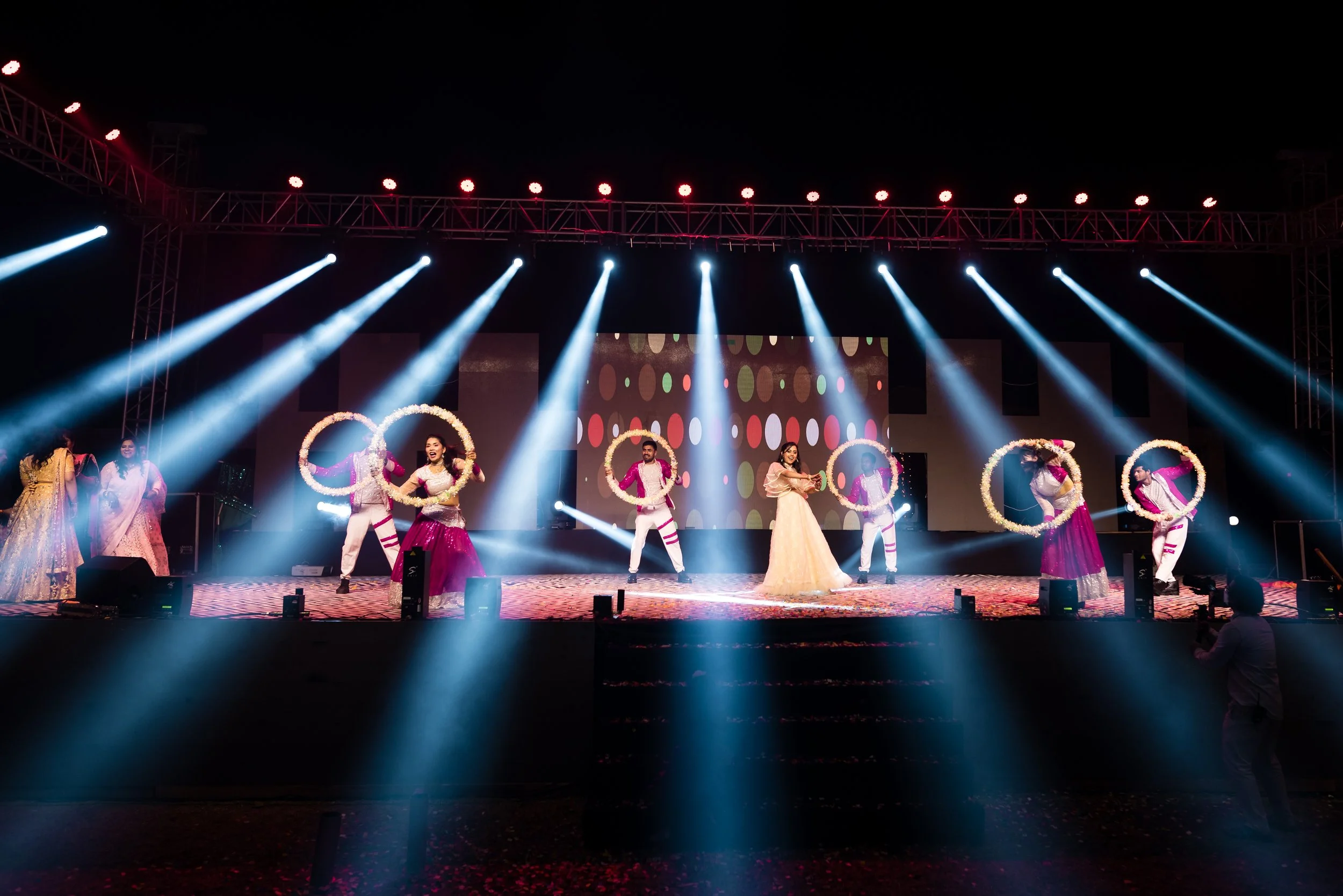 Performers dancing on a brightly lit stage at night, with some holding flower wreaths and a colorful polka dot backdrop.