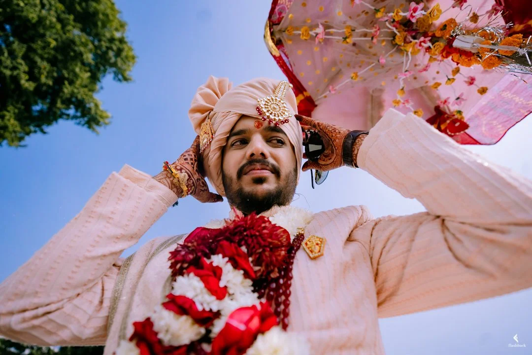 An Indian groom in traditional attire, adjusting his turban, with a floral umbrella in the background, during a wedding ceremony.