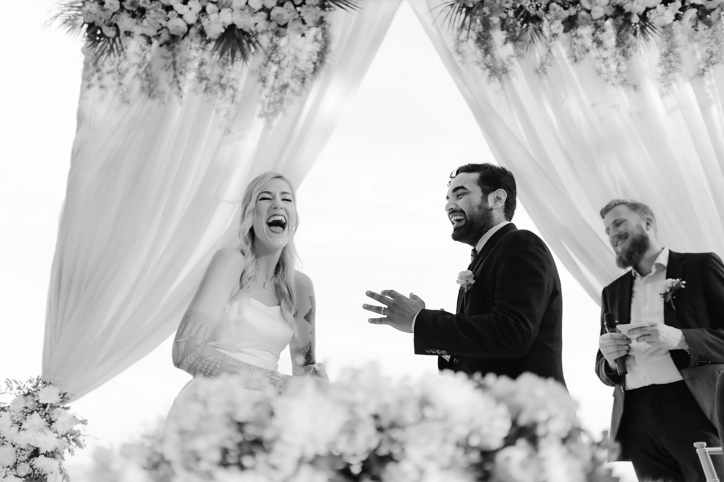 Black and white photo of a wedding ceremony, with a bride and groom laughing under a floral arch, and a man holding a microphone in the background.