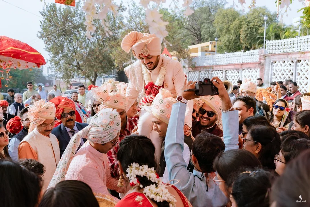 A traditional Indian wedding celebration with the groom being lifted on the shoulders of friends and family, surrounded by guests in colorful attire, outdoors with decorated umbrellas and floral decor.