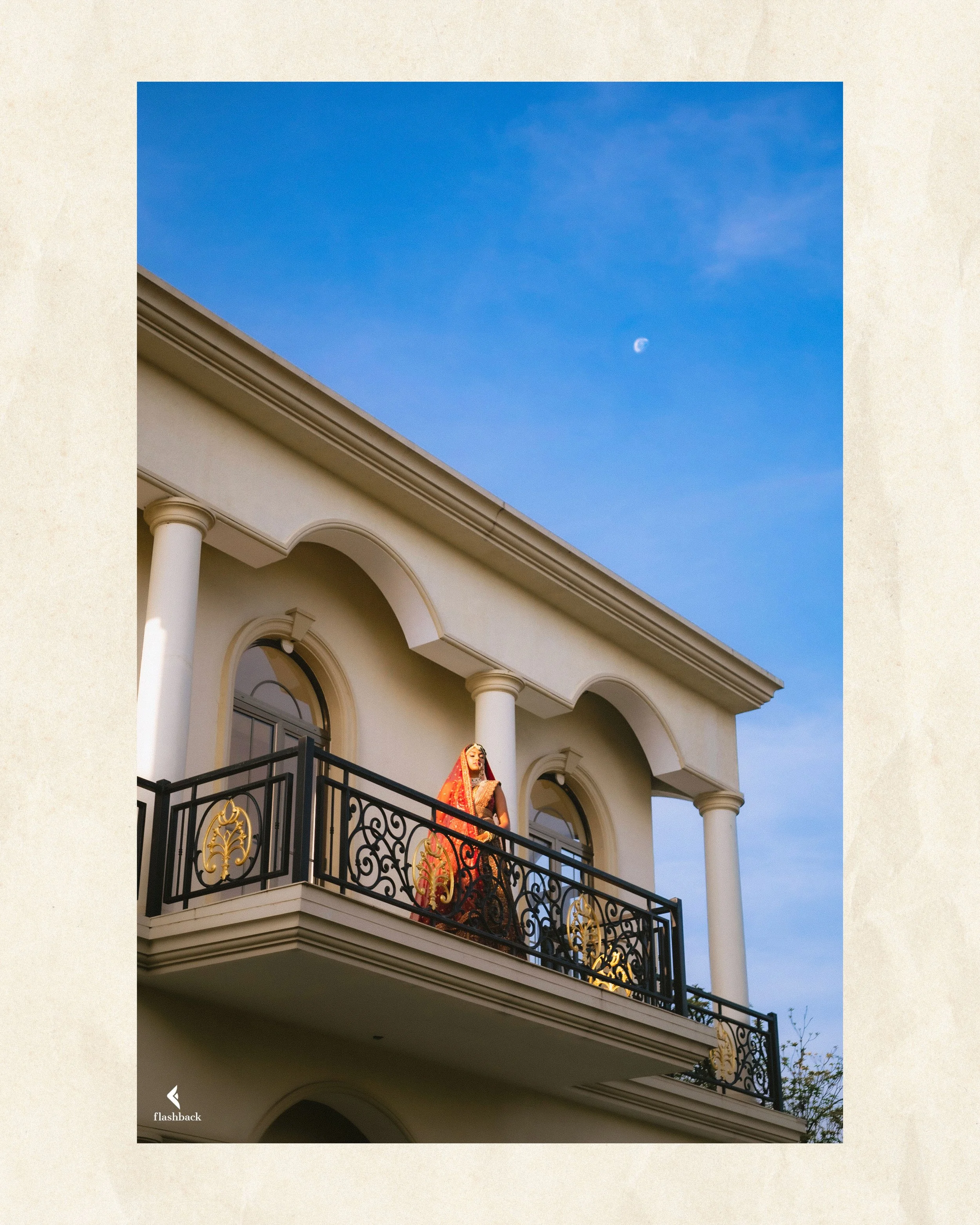 A woman in traditional Indian attire standing on a balcony of a white neoclassical building with columns, against a blue sky with a visible moon.