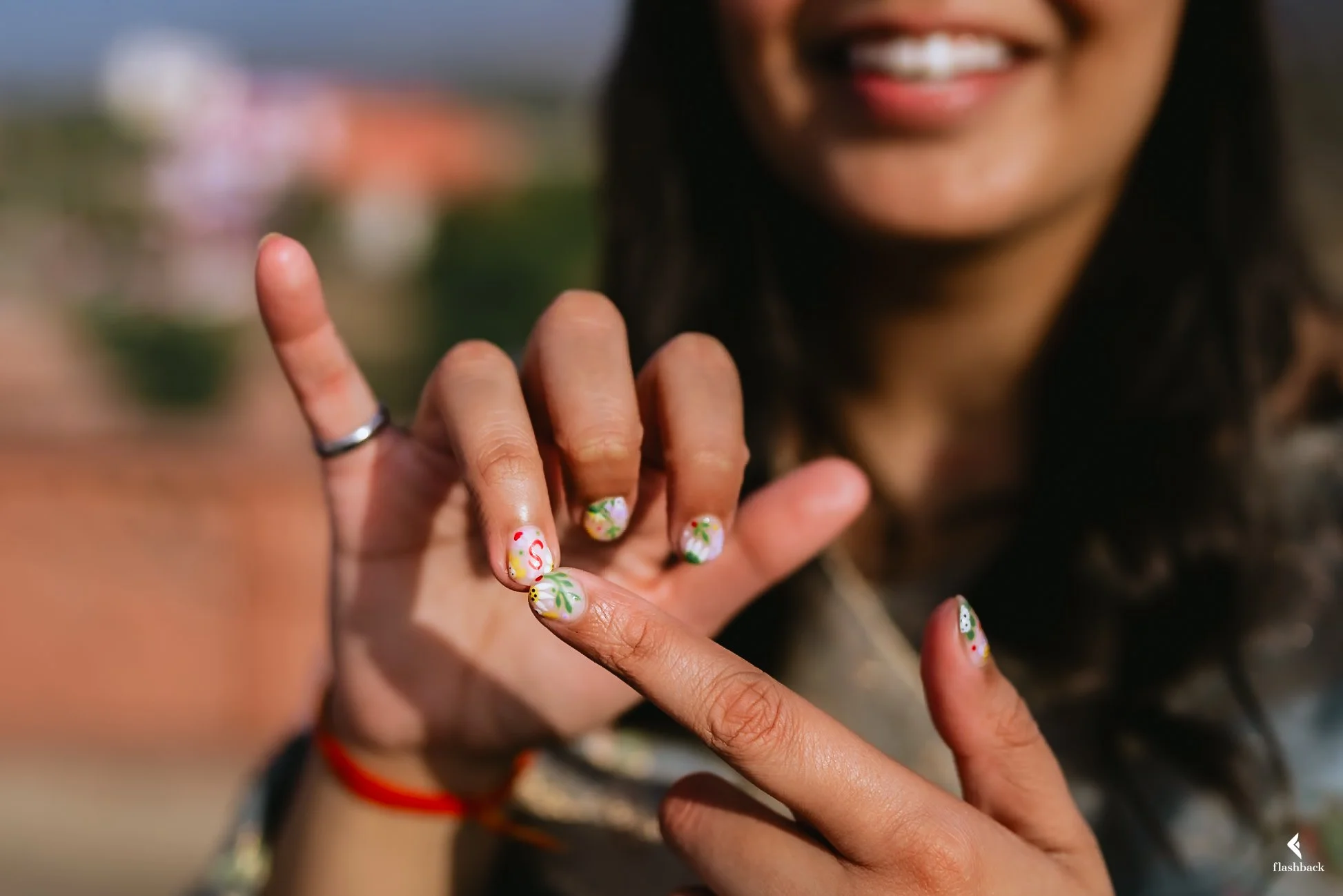 A woman with long dark hair showing her hand with decorated fingernails, smiling, with a blurred outdoor background.
