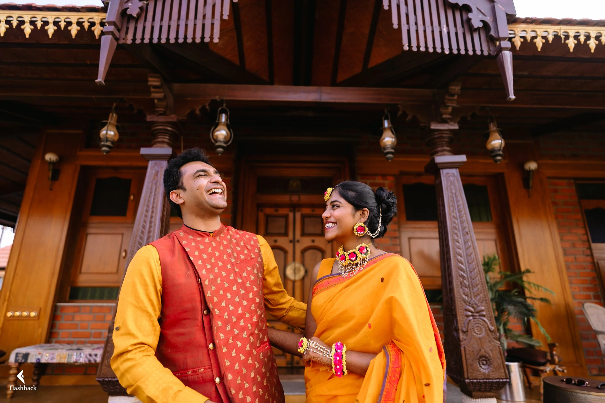 A joyful couple dressed in traditional Indian attire standing in front of a wooden house with ornate architecture, smiling and looking at each other.