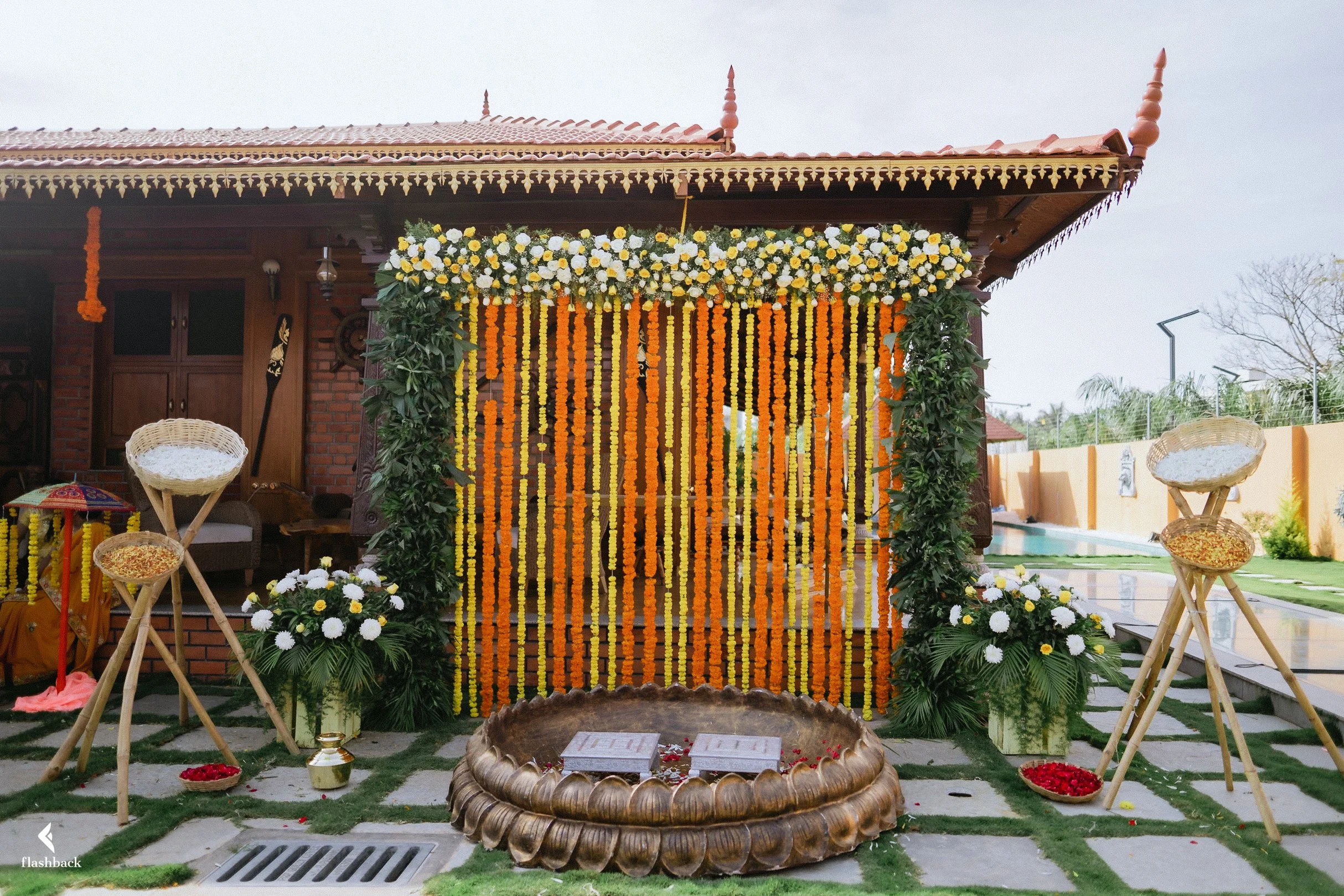 Decorated wedding mandap with orange and yellow marigold flower curtains, white and yellow floral arrangements, and traditional brass water pots, set outdoors near a pool.