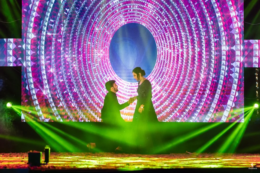 A couple on stage, with the man kneeling and holding the woman's hand, in front of a large, vibrant digital backdrop with pink and purple circular patterns, bright green stage lights shining.