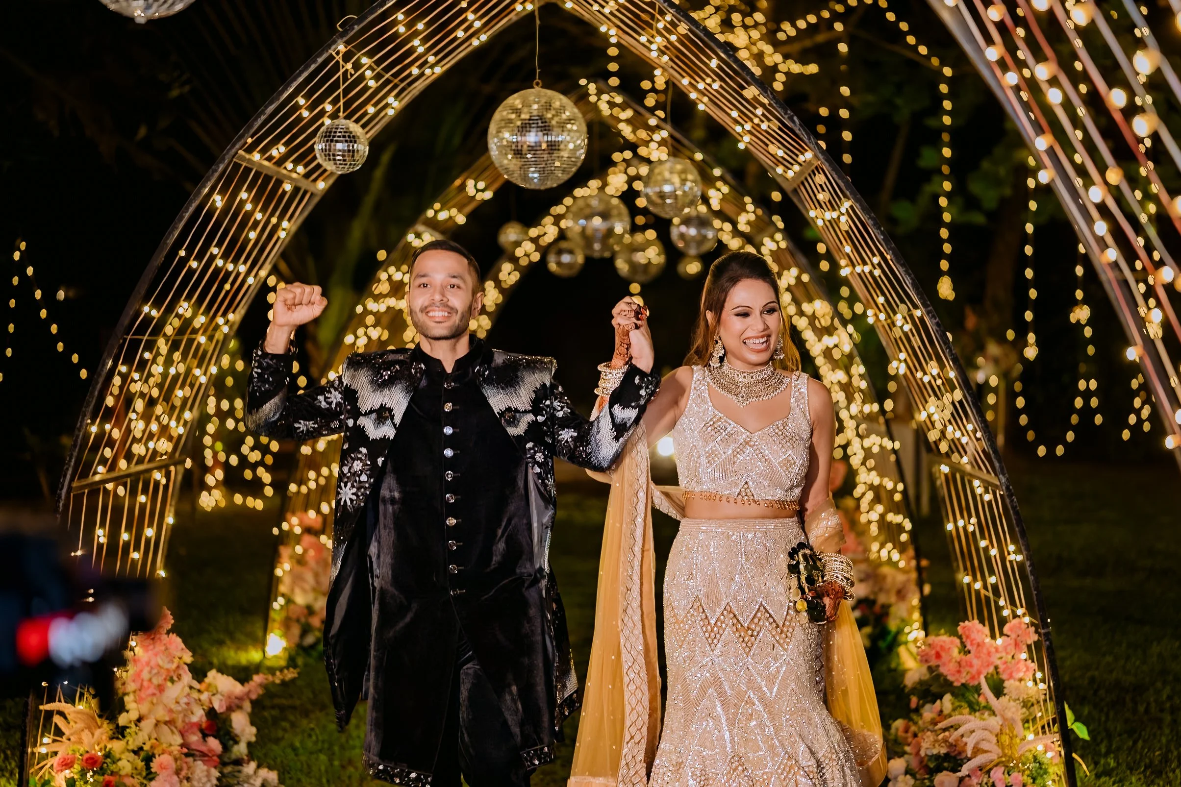 A couple celebrating their wedding at night under decorative arches with hanging disco balls and string lights, surrounded by floral arrangements.