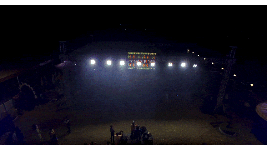Nighttime outdoor stage with bright lights, a colorful screen, and people gathered on the ground.