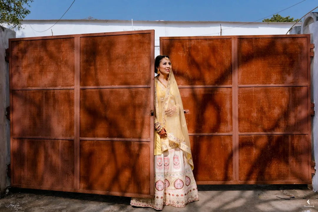 A woman in traditional Indian attire standing beside a large, rusty metal gate, partially hidden behind it, with shadows of tree branches cast on the gate.