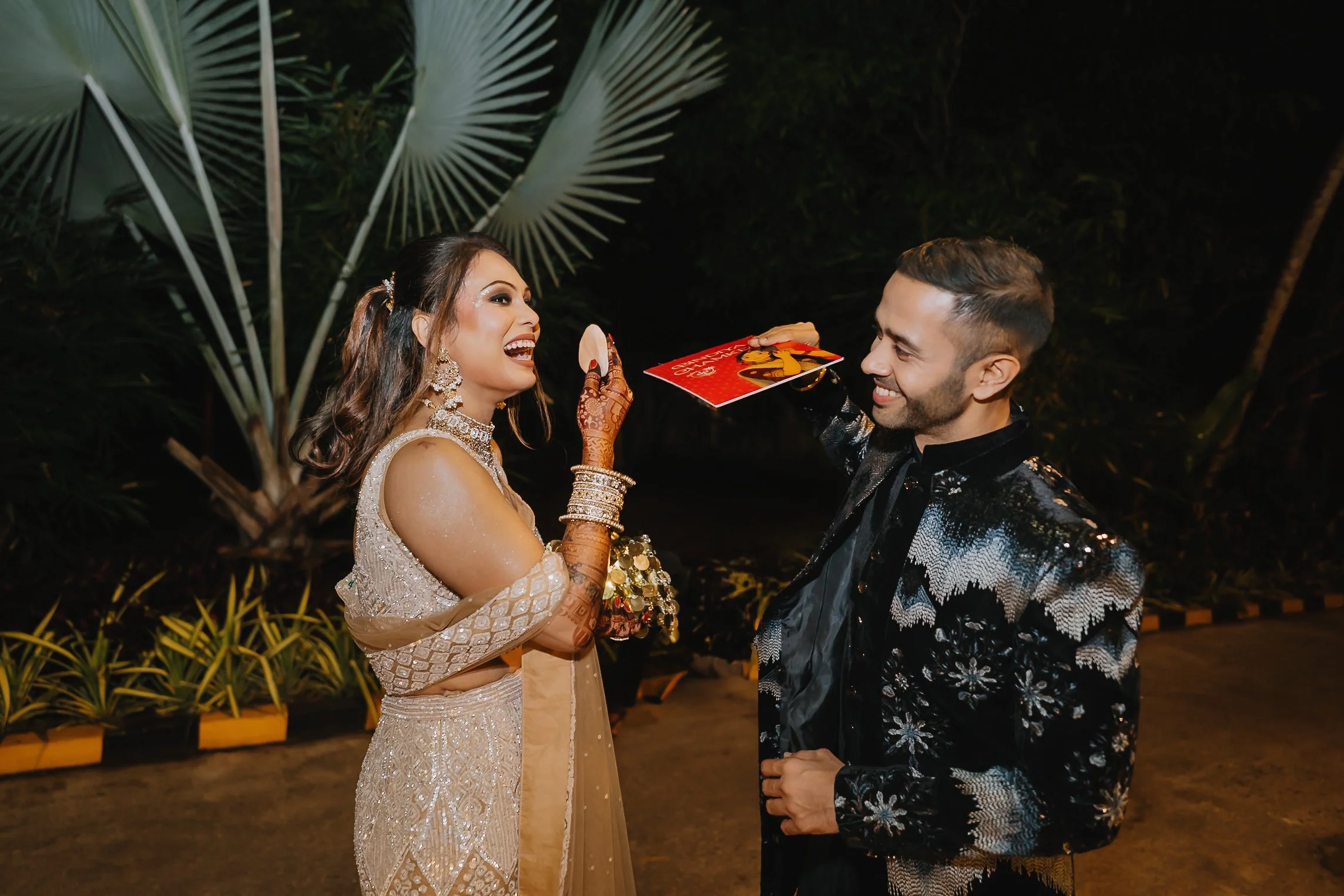A woman dressed in traditional Indian attire with jewelry and henna on her hands, smiling and holding a decorated item, standing next to a man in a black patterned blazer, both engaged in a joyful moment outdoors at night surrounded by green plants.