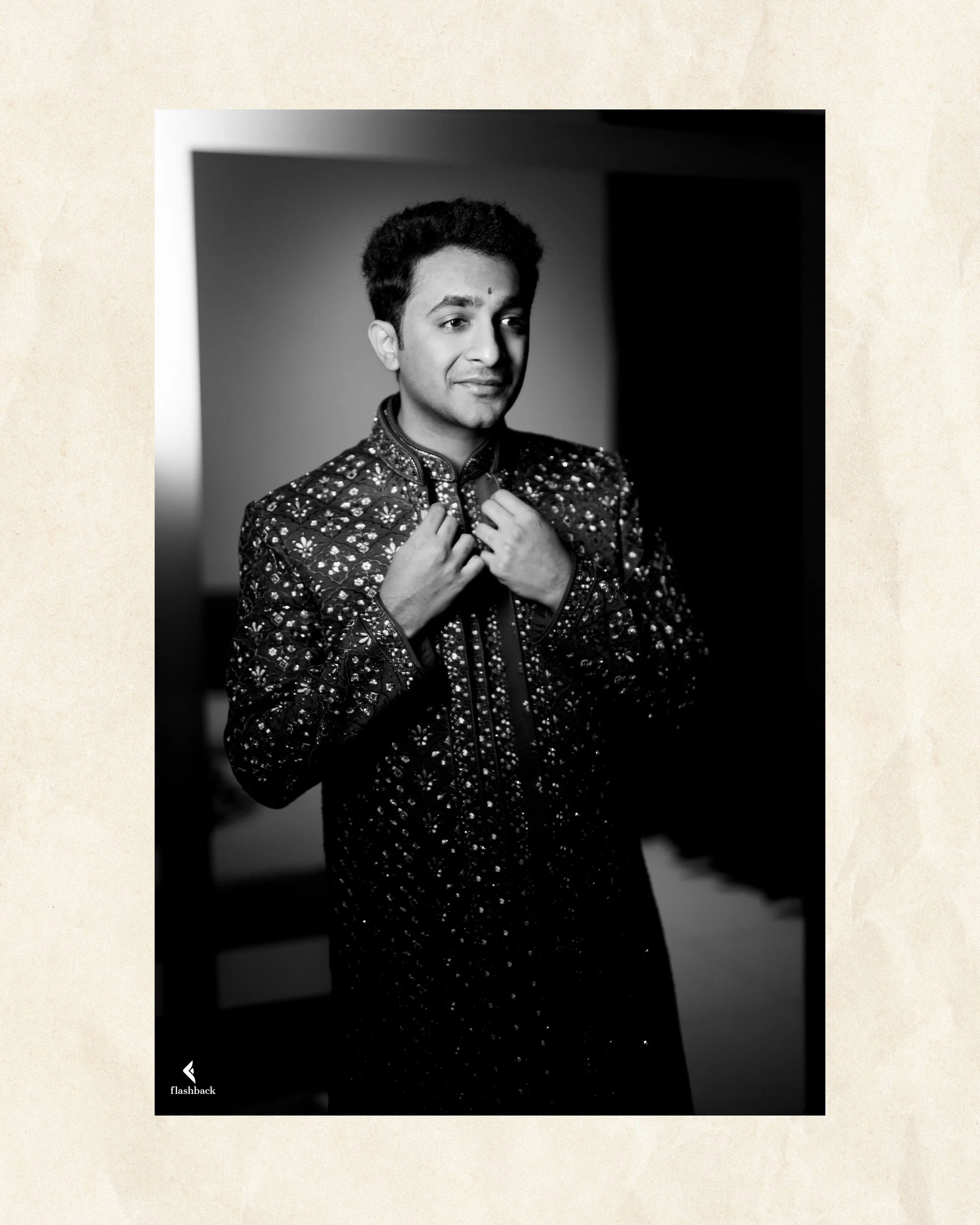 A young man in traditional Indian attire, adjusting his embroidered outfit in a black and white photograph.