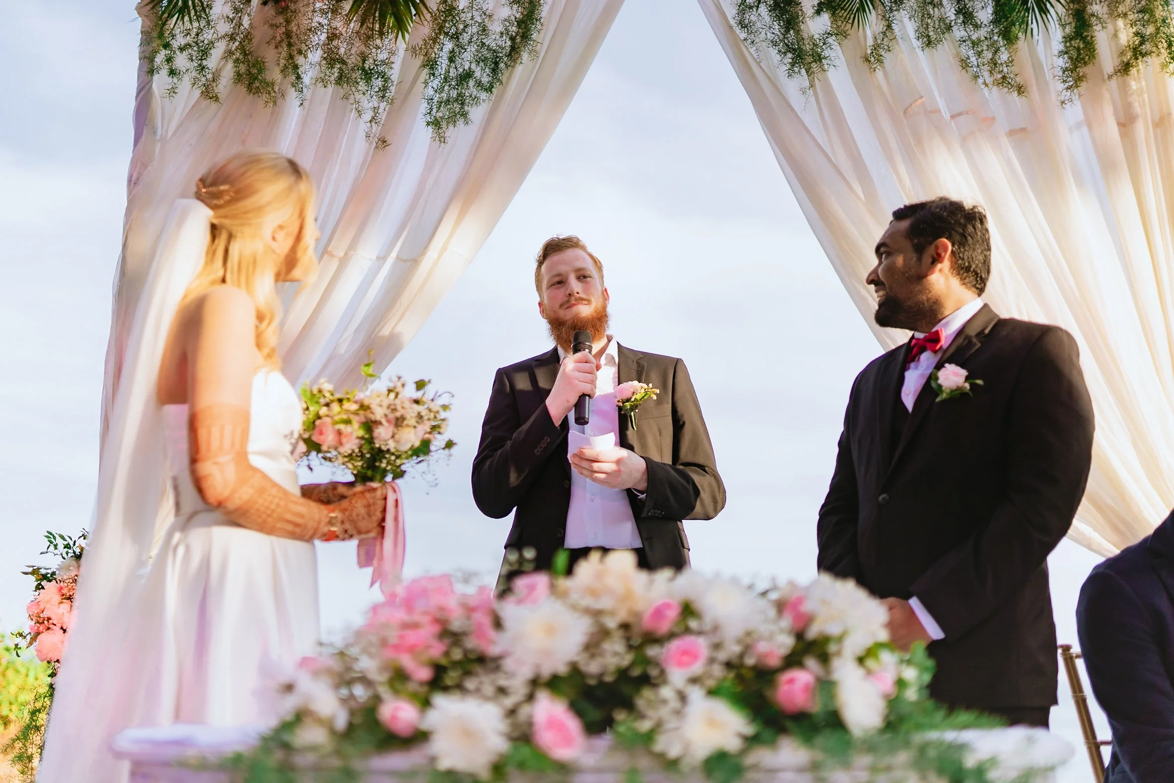 A wedding ceremony outdoors with a bride and groom standing under a decorated arch, while a man with a microphone officiates. The bride has long blonde hair and wears a white dress with lace sleeves, holding a bouquet of pink and white flowers. The g