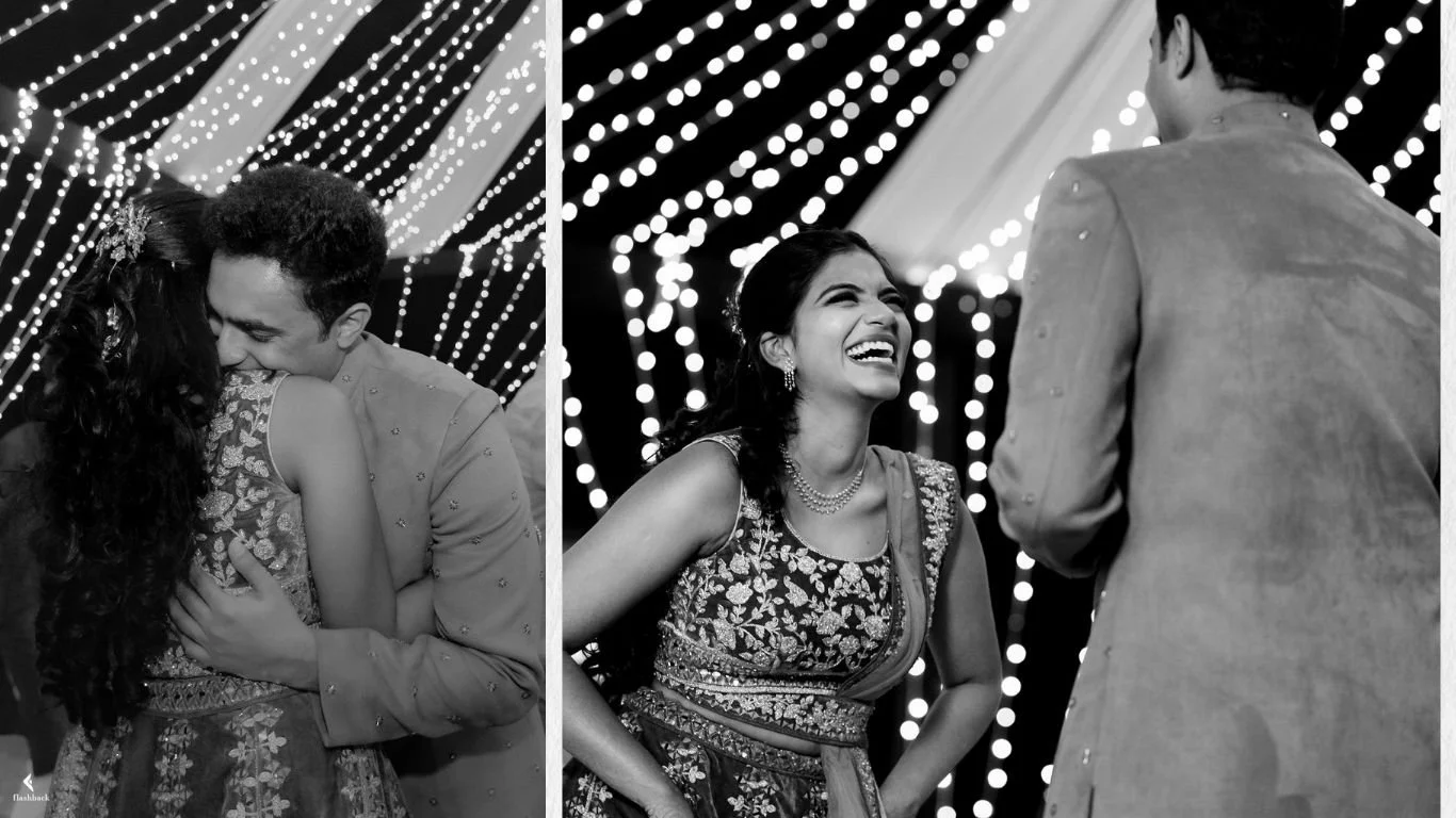 A black and white photo featuring a joyful woman dressed in traditional Indian attire, laughing as she interacts with a man in a kurta at a festively decorated event with string lights in the background.