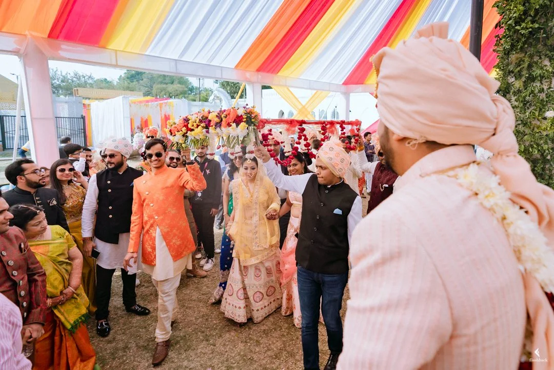 People dressed in traditional Indian clothing at a wedding celebration, with some holding flower arrangements and a floral backdrop, under a colorful tent canopy.