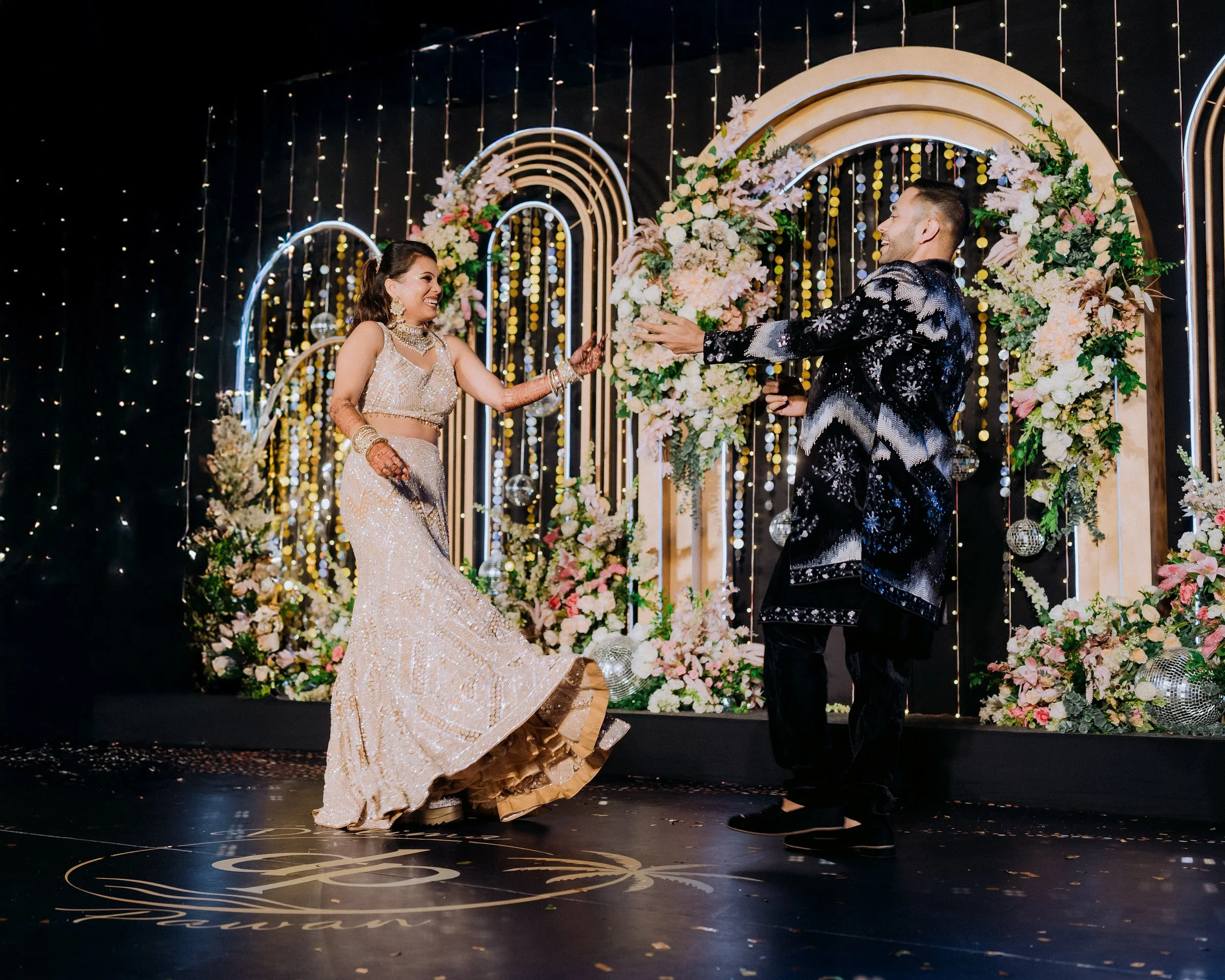 A bride and groom are dancing at their wedding reception, surrounded by floral arrangements and decorative arches with string lights in the background.