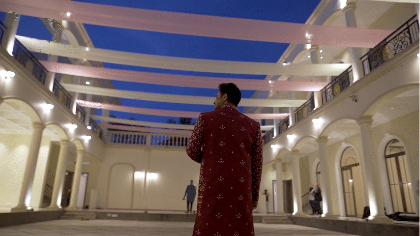 Man wearing a red patterned coat standing in a courtyard at night with white columns, arches, and pink and white fabric drapes overhead.