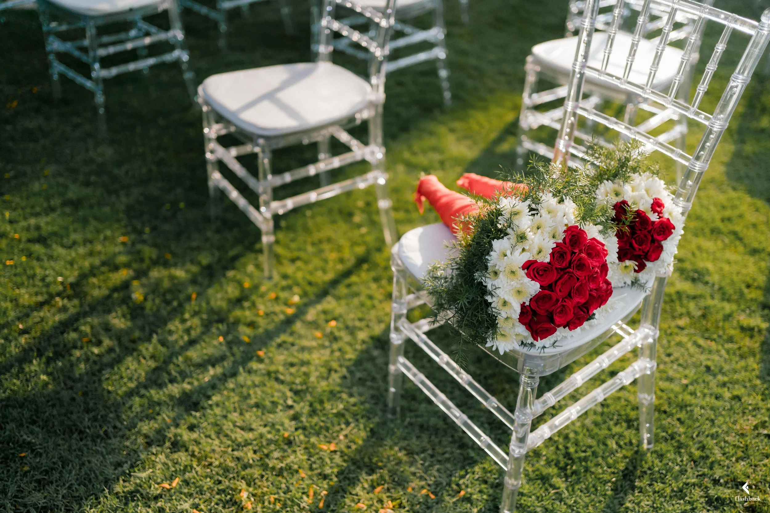 A clear acrylic wedding chair with a bouquet of red roses and white flowers attached to the back, set outdoors on a grassy lawn with other similar chairs nearby.