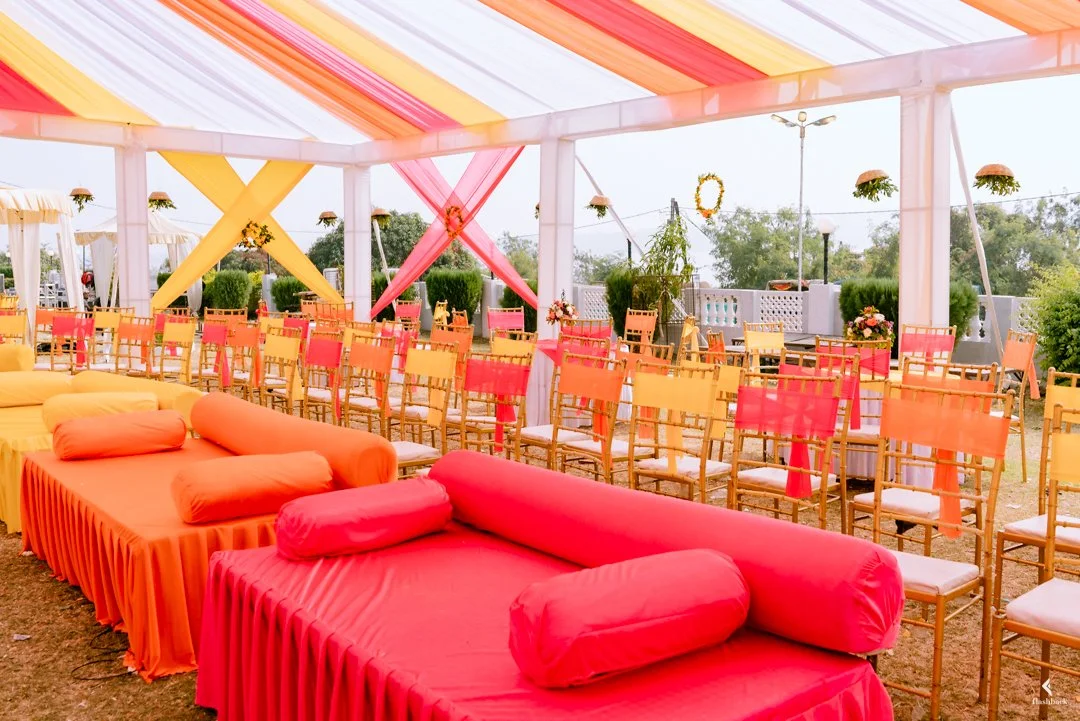 Colorful wedding or event tent with pink, yellow, and orange drapes, rows of yellow and pink chairs, and decorated seating area with pink and orange cushions and bolsters.