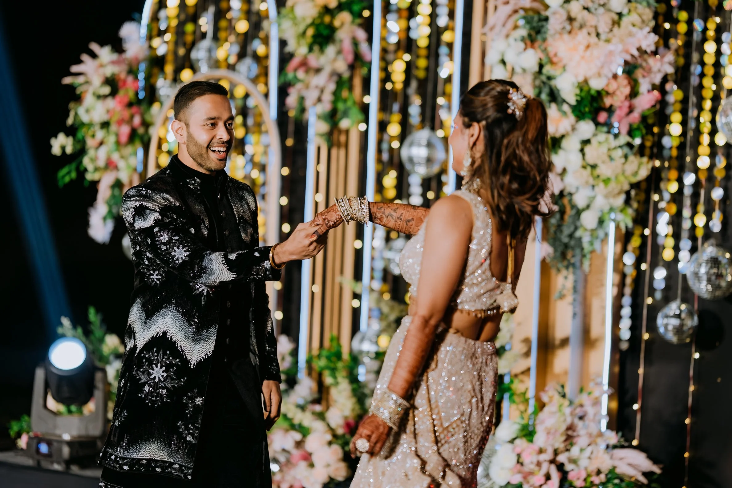 A couple at a wedding, the groom smiling as he holds the bride's hand, standing in front of a floral backdrop with lights.
