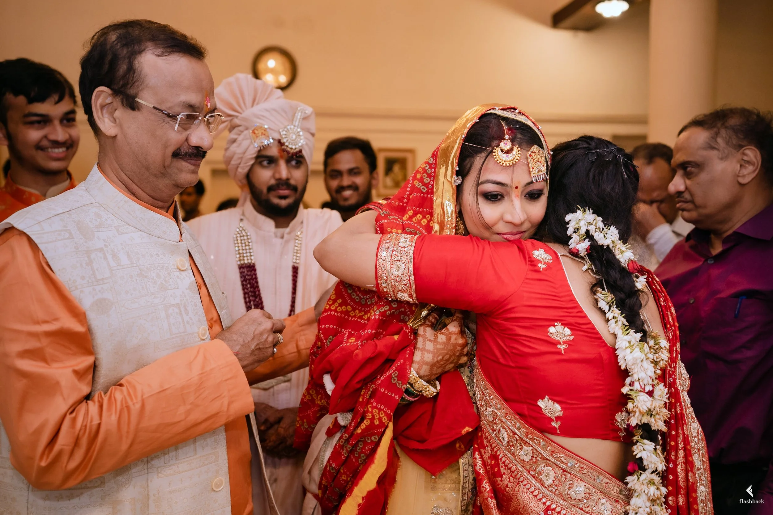 Indian bride hugging her mother at a wedding celebration surrounded by smiling guests in traditional attire.