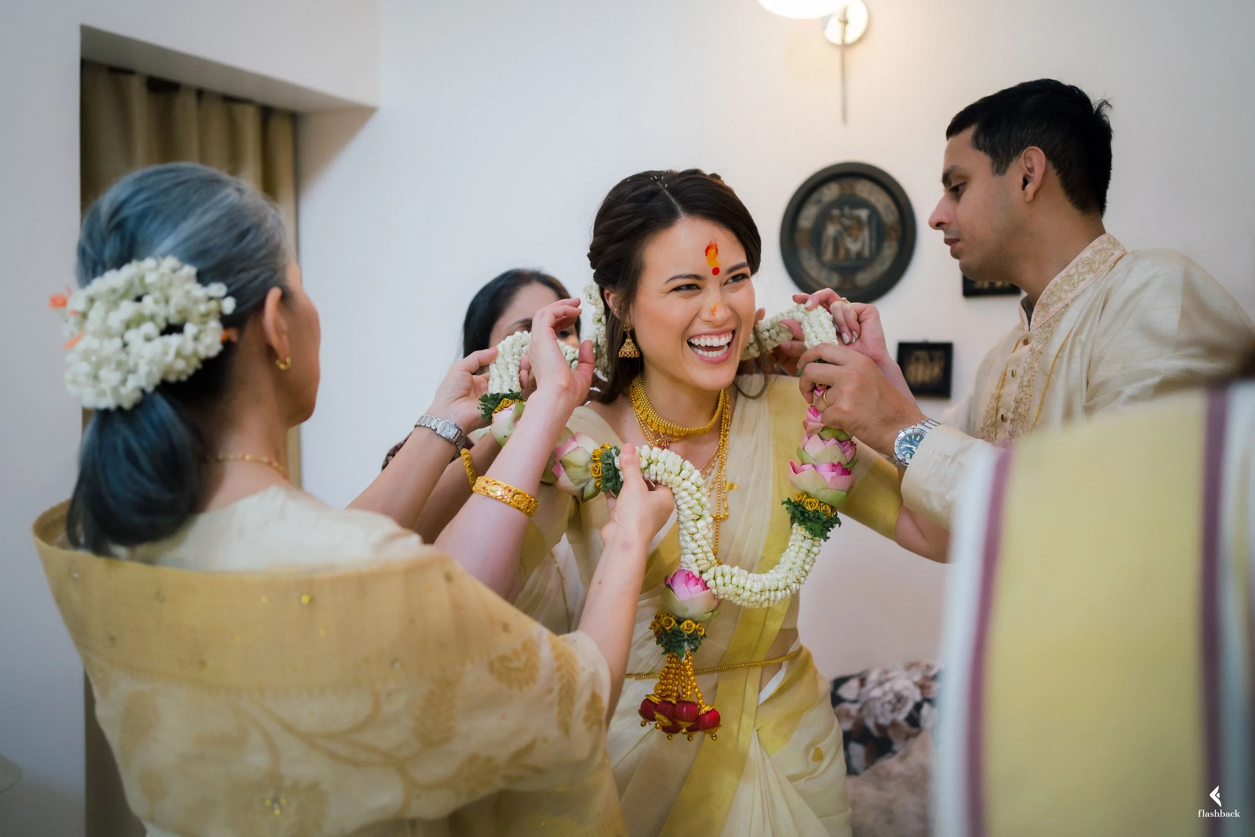A woman in traditional Indian attire smiling as she is adorned with flower garlands by women and a man in a ceremonial setting.