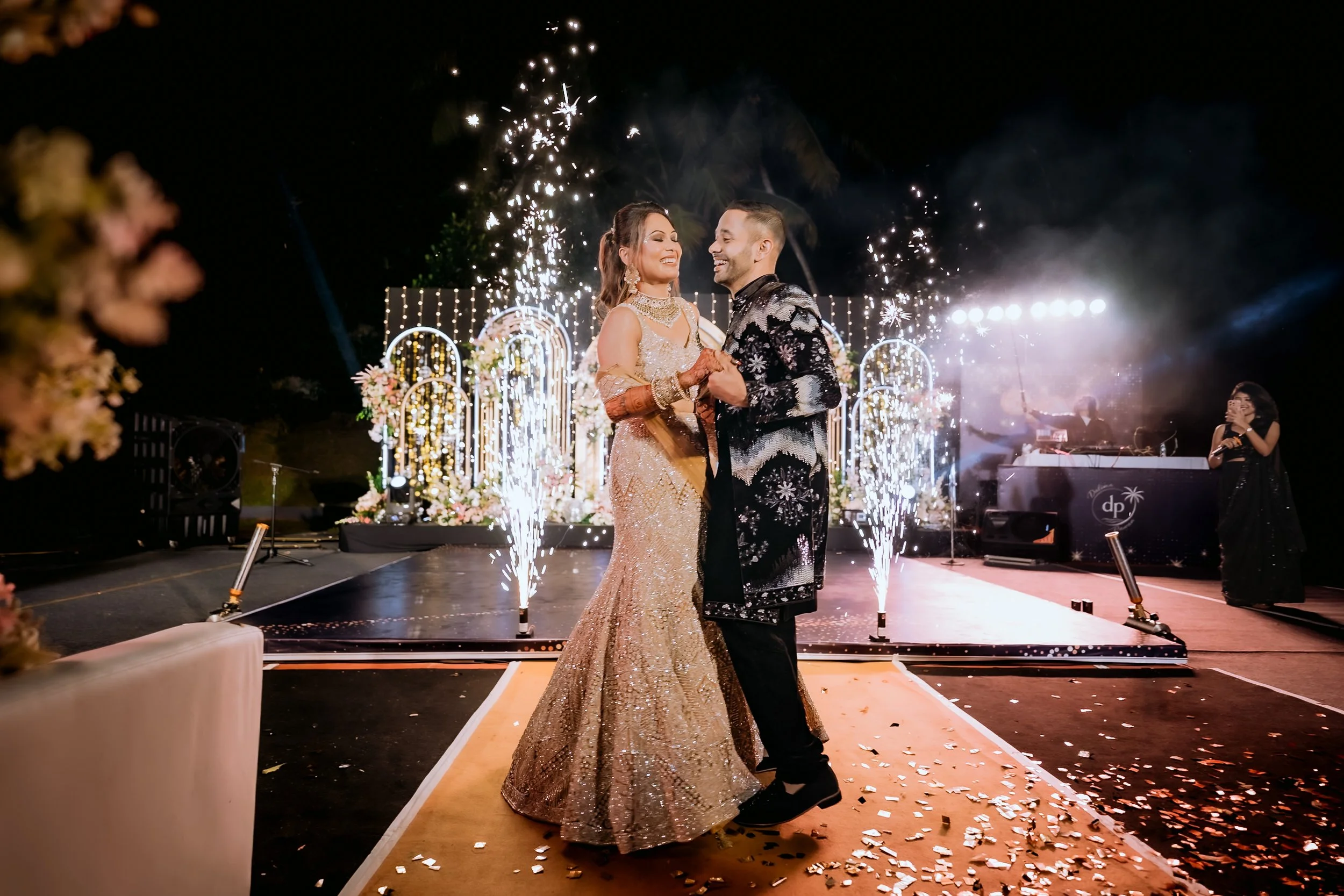 A bride and groom dancing during their wedding celebration at night, with sparklers and fireworks in the background, decorated with flowers and string lights.