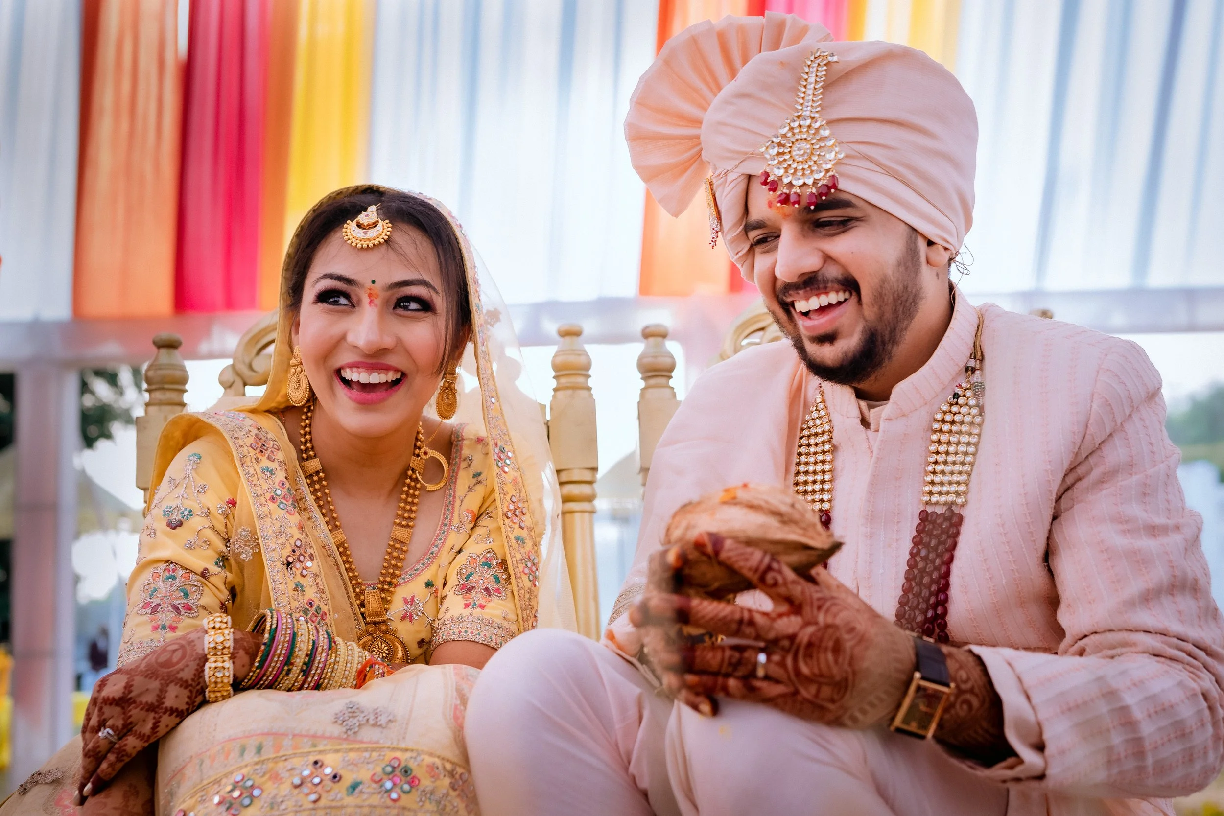 Indian wedding couple dressed in traditional attire smiling and sitting together during the ceremony, with colorful drapes in the background.