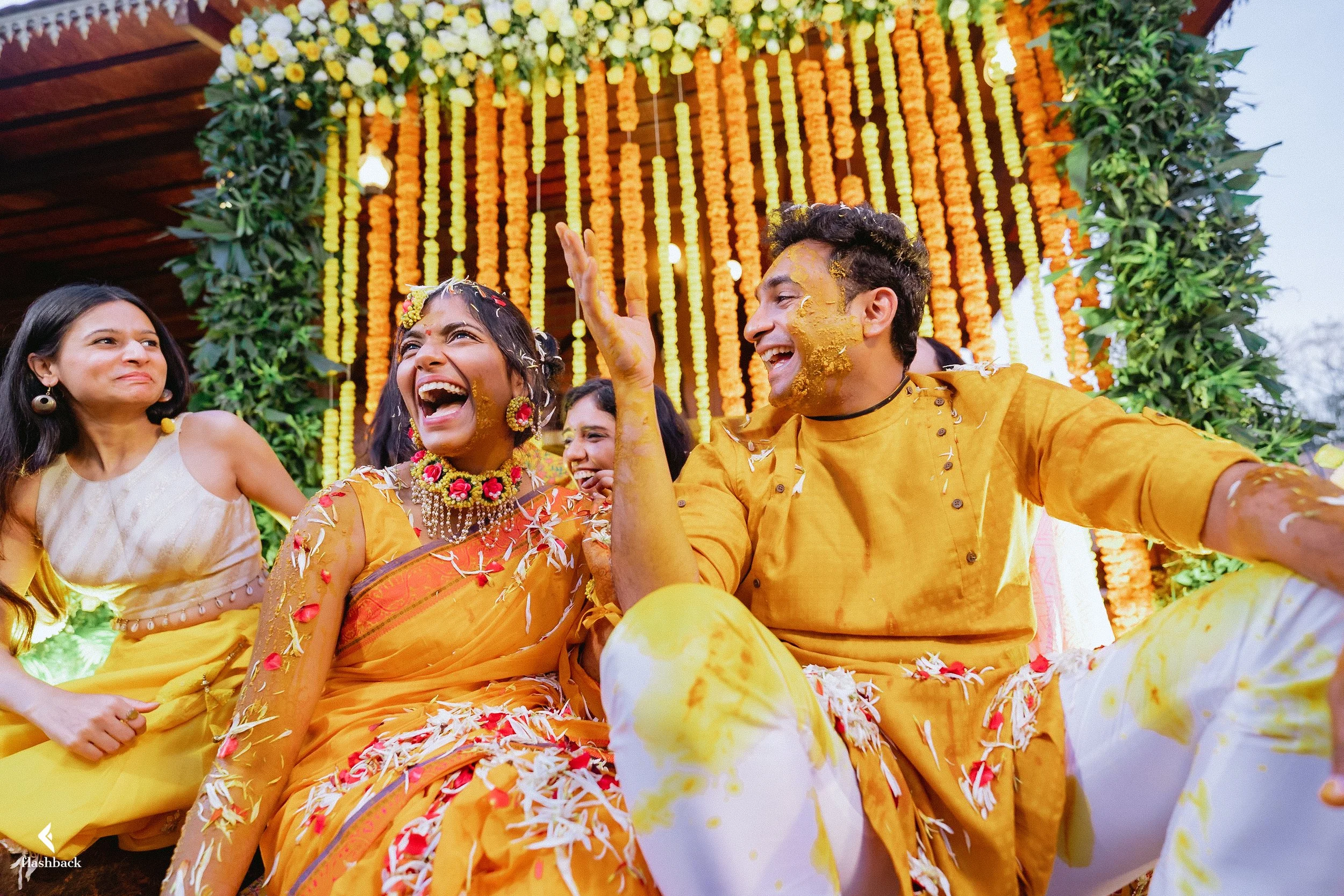 Celebrating a traditional Indian wedding with women and men dressed in yellow and orange clothing, surrounded by flower decorations, and smiling joyfully.
