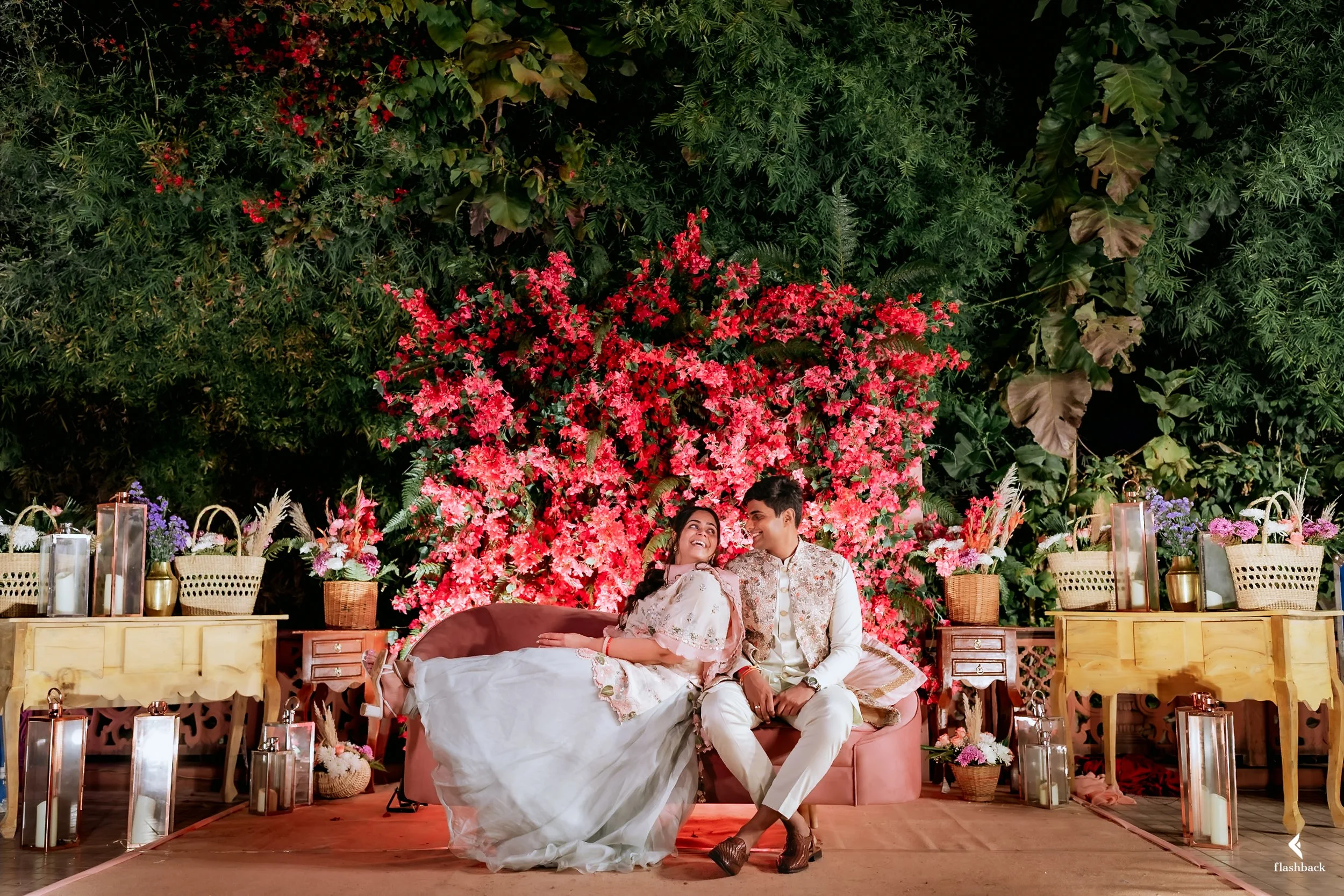 A bride and groom sitting on a pink sofa in front of a vibrant floral backdrop with pink and red flowers. The setting is decorated with lanterns, candles, baskets, and vases filled with flowers, creating an elegant and romantic ambiance.
