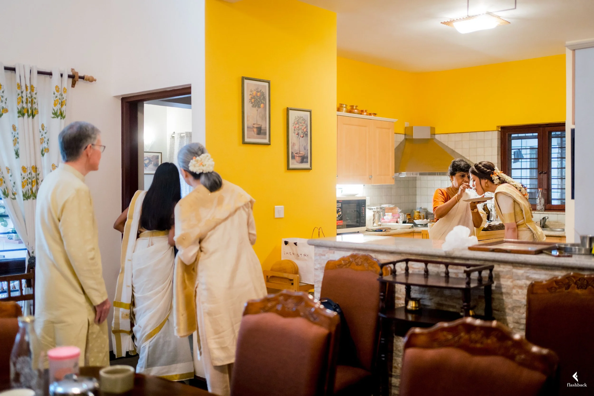 Family gathering at home with several women wearing traditional Indian sarees, a man is present, and women are sharing a meal in the kitchen.