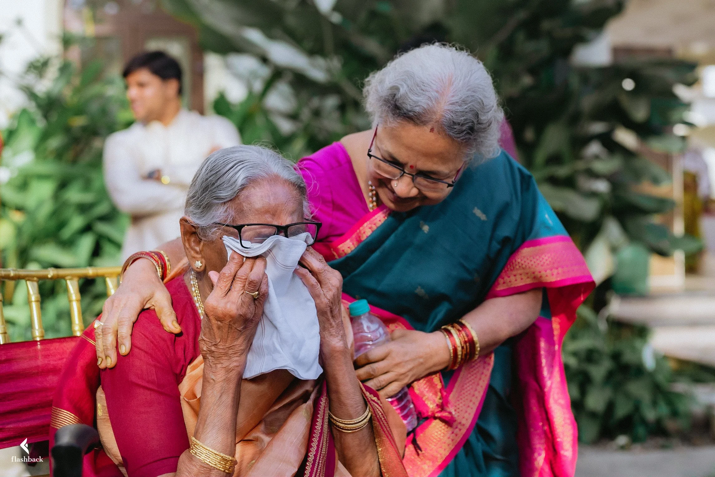 An elderly woman, wearing glasses and traditional Indian clothing, is emotional and wiping tears with a tissue. A woman beside her, also in traditional Indian attire, is comforting her with a smile. They are outdoors with greenery in the background.
