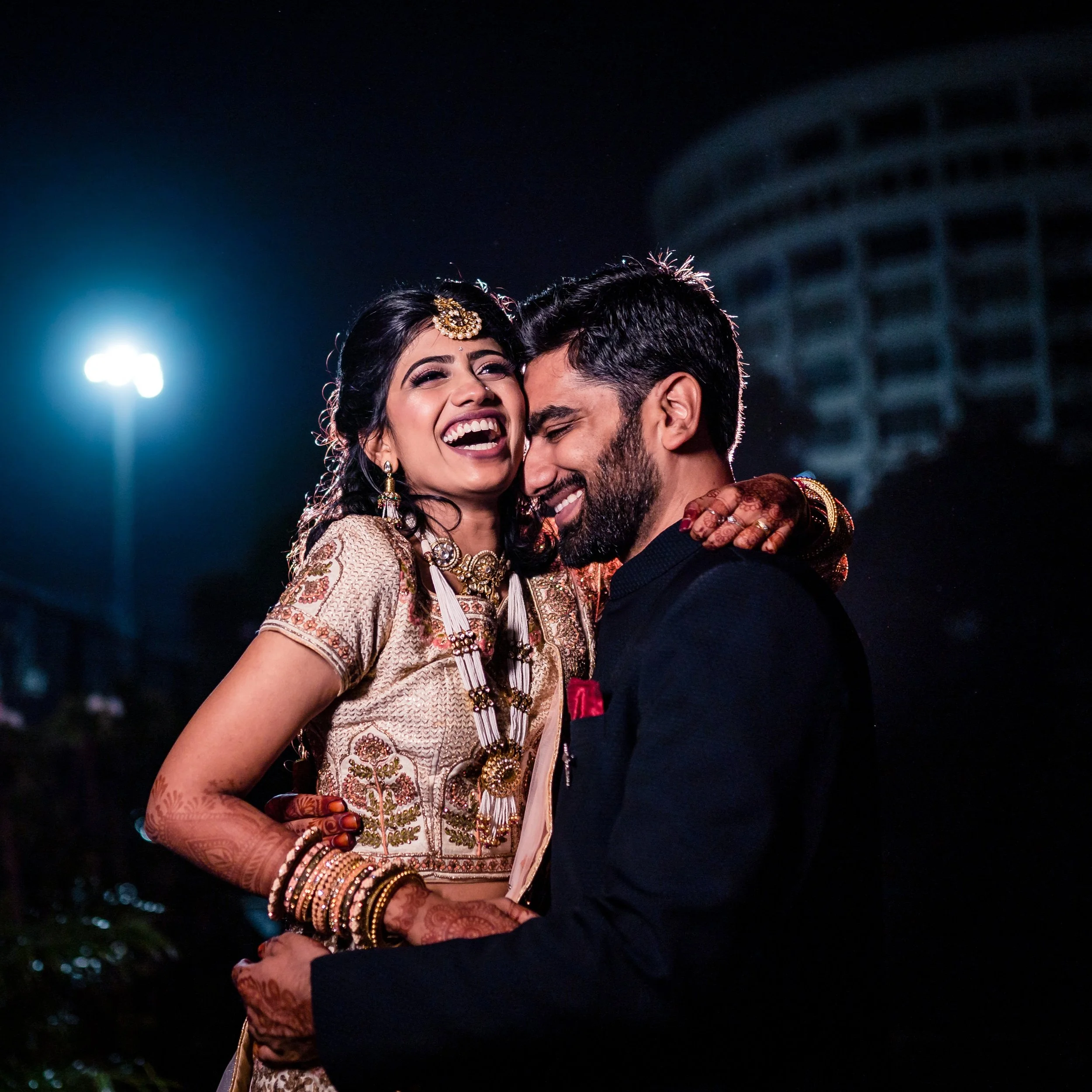 A joyful couple in traditional Indian wedding attire sharing a happy moment at night.