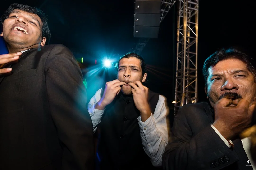 Three men at a party or event, making funny faces, with colorful lights and a stage or rigging in the background.