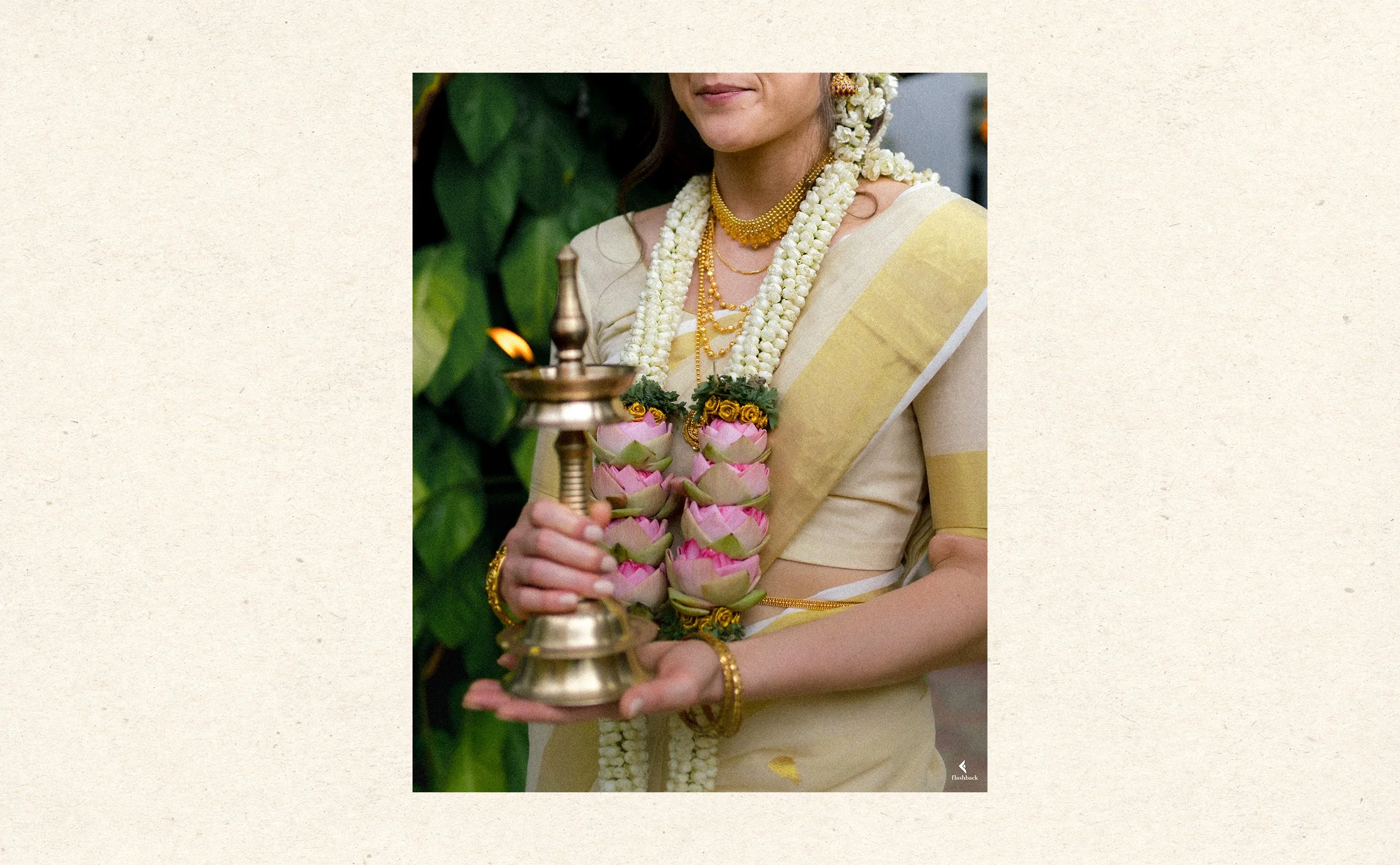 Woman in traditional Indian attire holding a brass lamp, adorned with multiple flower garlands, standing in front of green foliage.