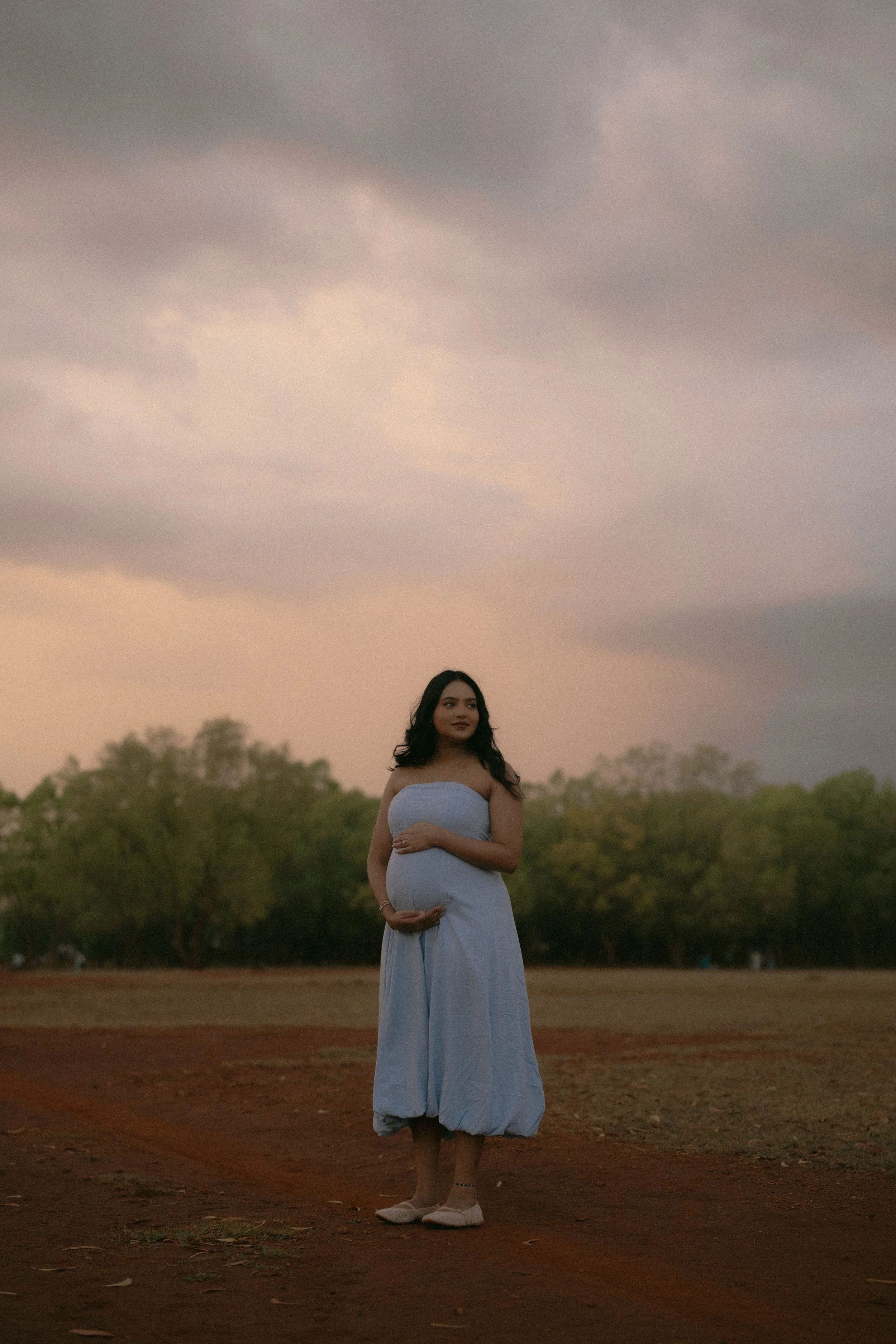 Expecting mother standing alone in a field beneath a pink and grey storm sky