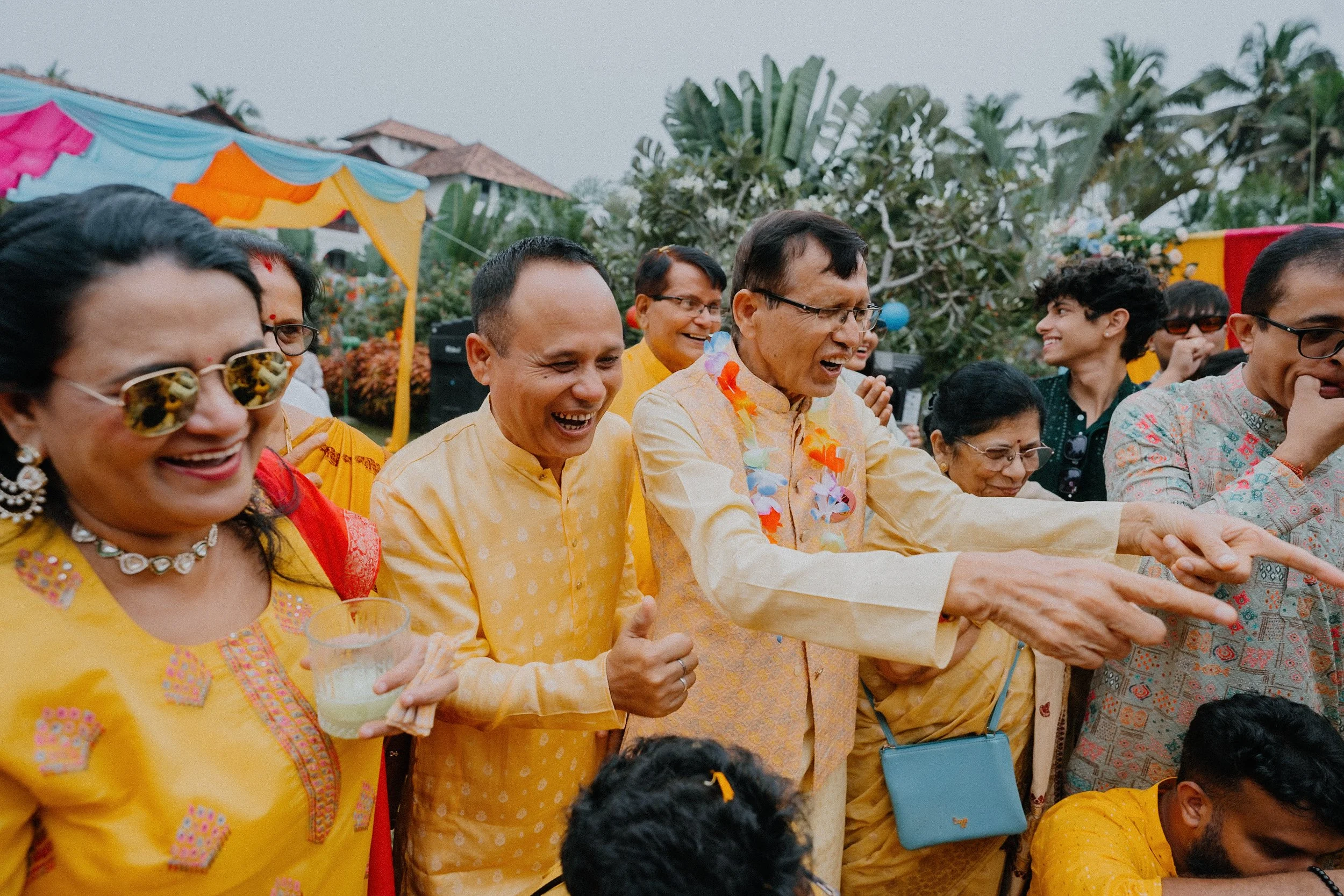 People celebrating at an outdoor event, wearing traditional Indian clothing, with colorful decorations and greenery in the background.