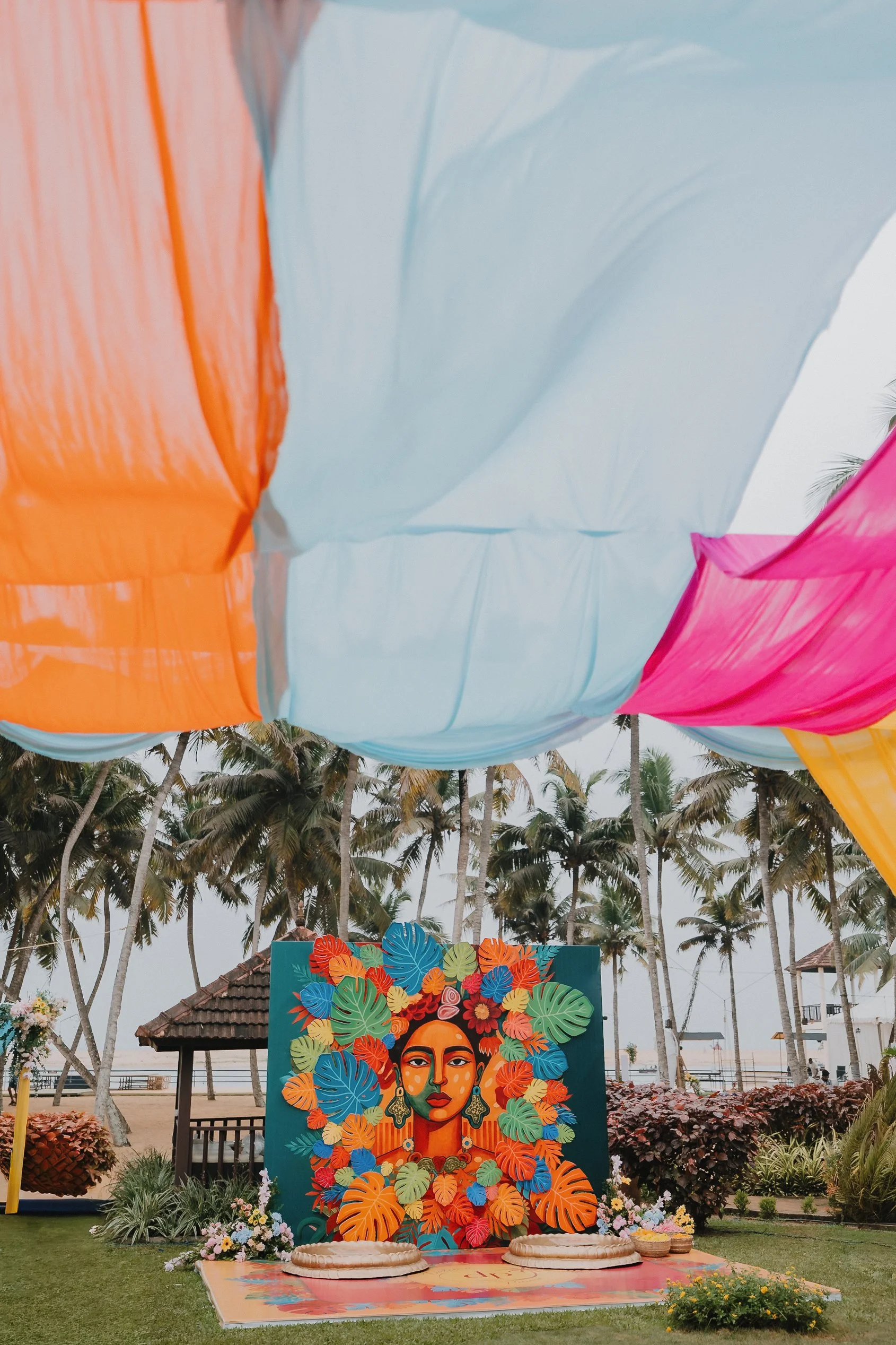 A colorful outdoor art display featuring a large portrait of a woman with flowers and leaves in her hair, set against a backdrop of palm trees and cloudy sky.