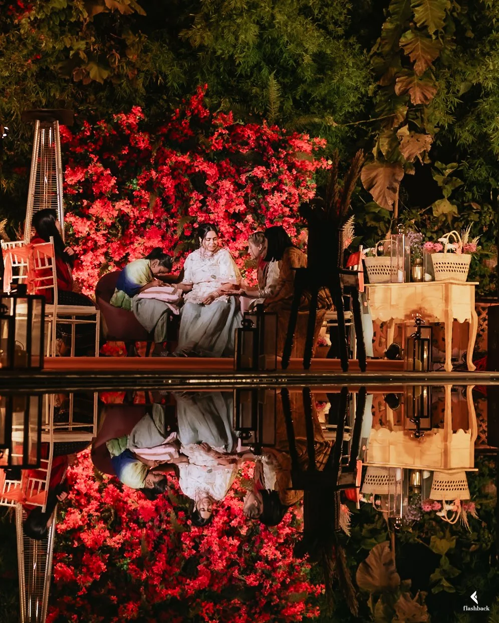 People sitting together on a stage decorated with red flowers and greenery, with a mirror-like reflection of the scene below.