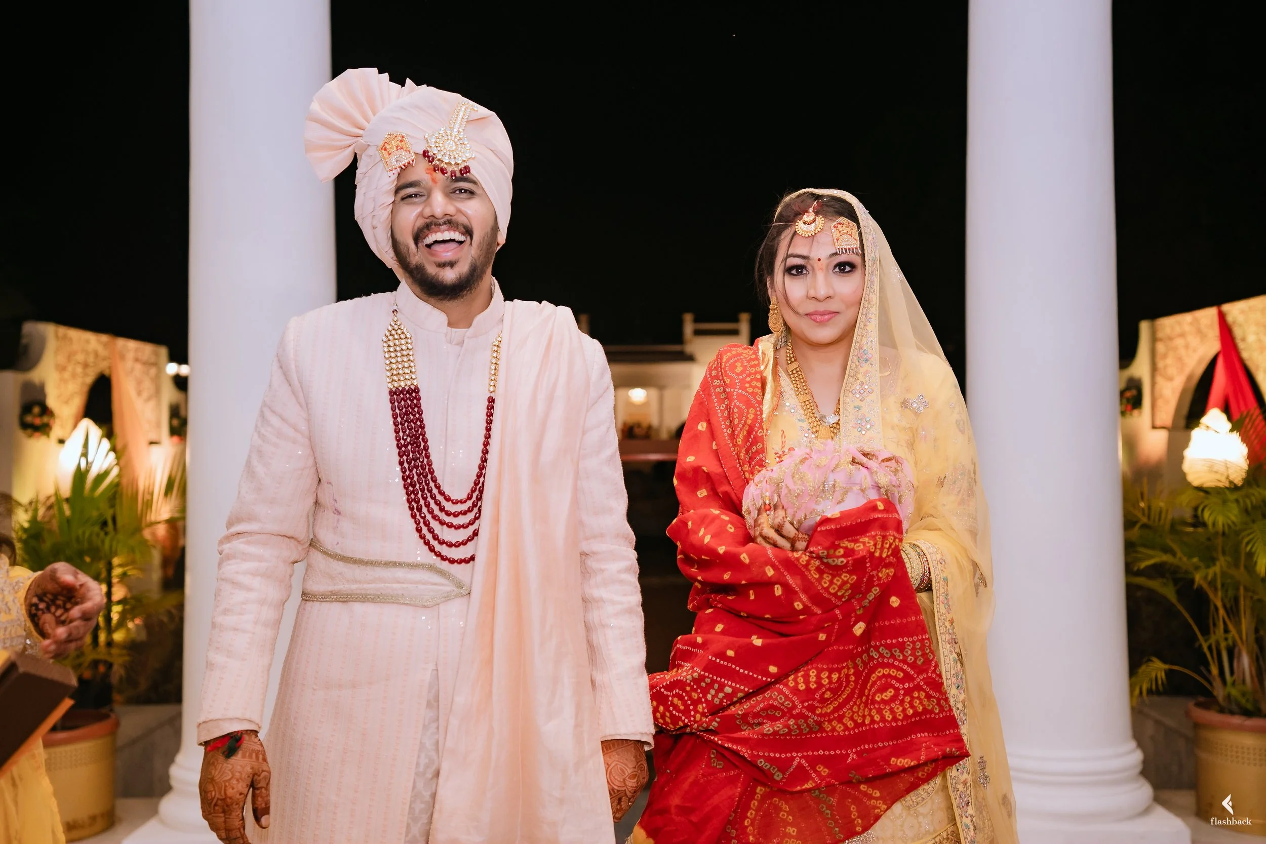 A couple dressed in traditional Indian wedding attire, standing in front of white pillars at night. The groom is wearing a cream-colored sherwani with red necklaces and a pink turban. The bride is wearing a red and gold saree with a yellow dupatta, a