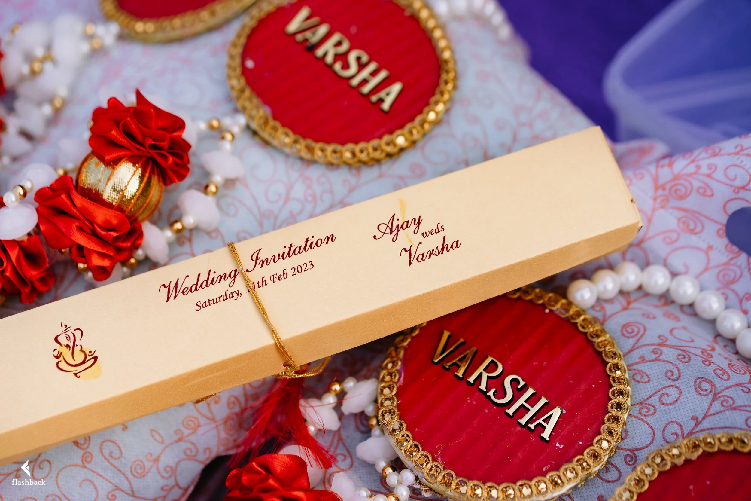 Wedding invitation card with the names Ajay and Vansha, dated Saturday, February 11th, 2023, surrounded by red and gold decorative items, jewelry, and embroidered fabric.