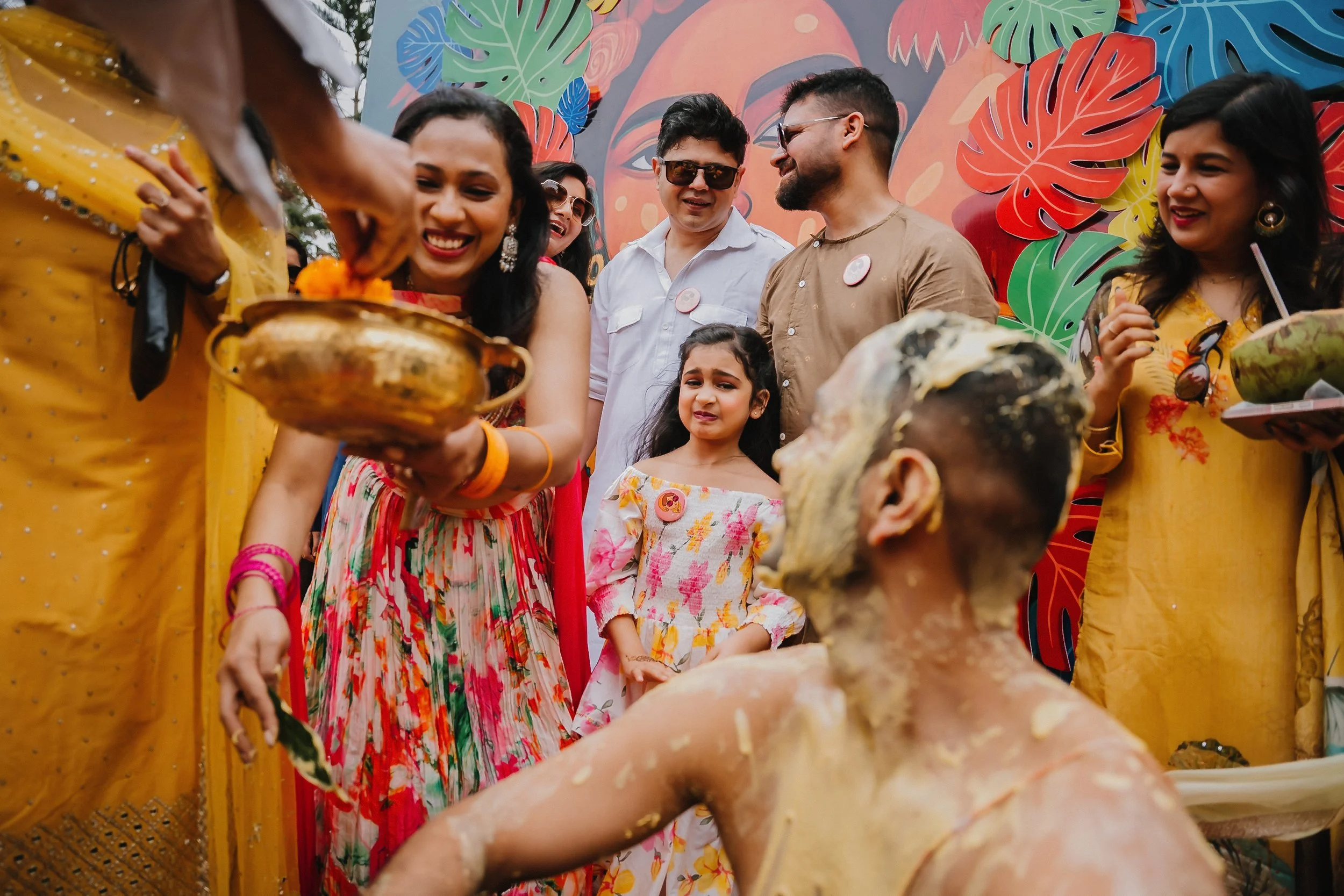 A group of people celebrating a traditional Indian ritual, with one person holding a brass pot with marigold flowers, while others gather around smiling and wearing colorful attire, in front of a vibrant mural with tropical leaves.