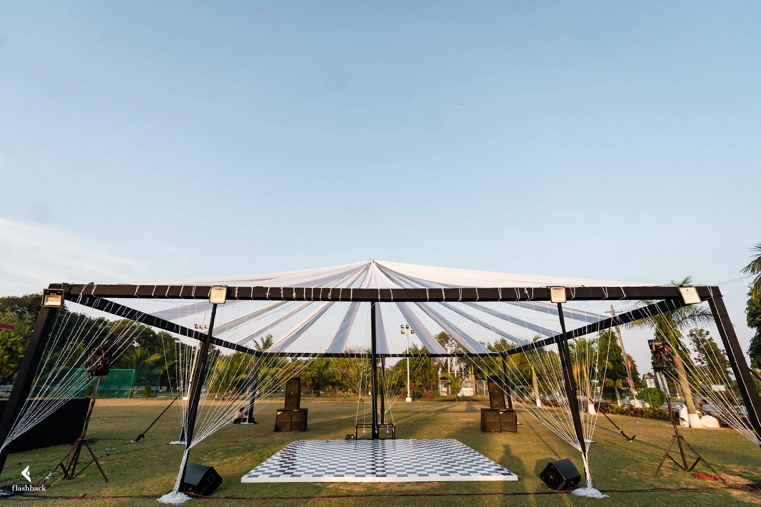 Outdoor stage setup with a black and white checkered floor, covered by a white tent-like canopy, surrounded by trees and open sky.