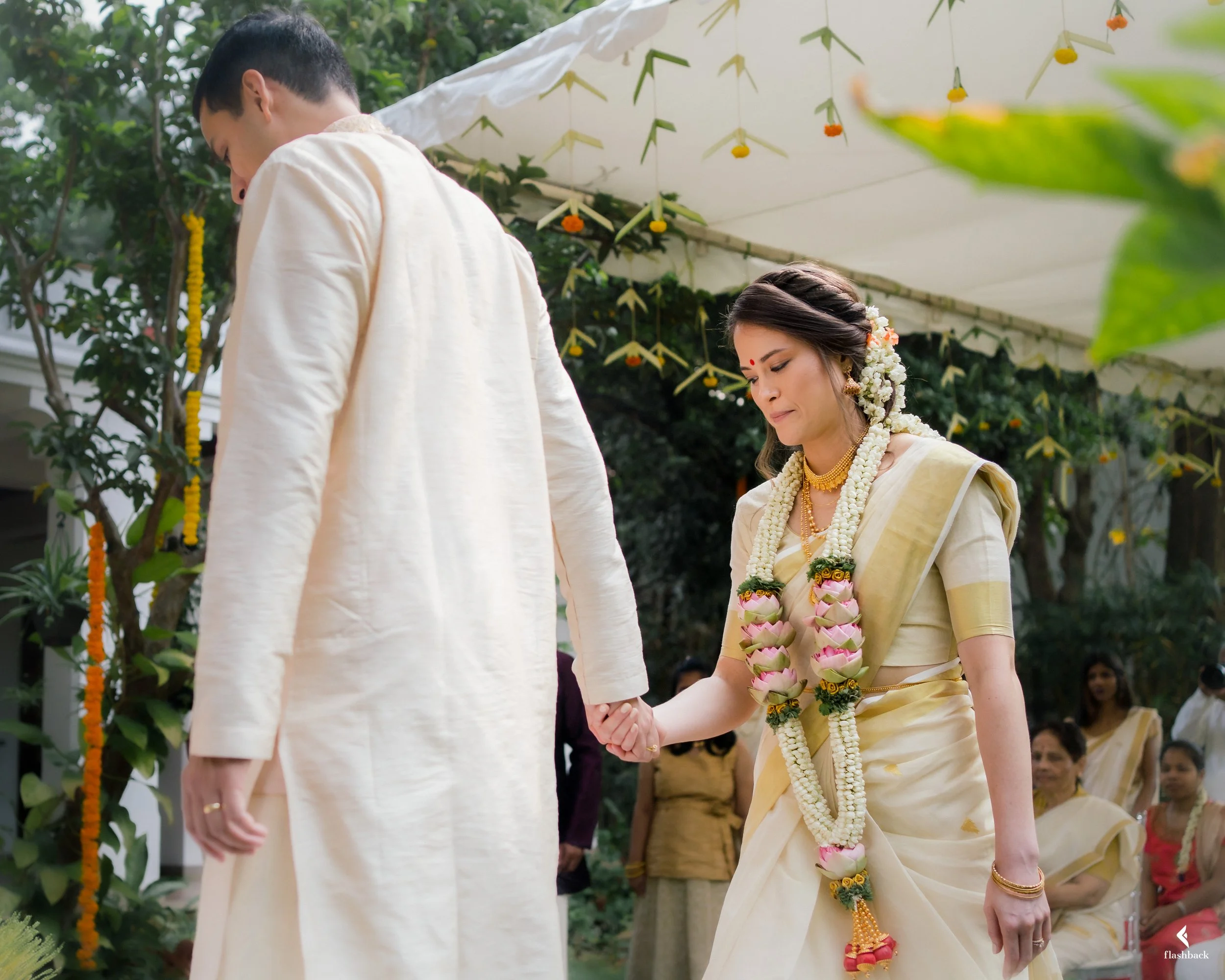 A woman in a traditional cream-colored saree with floral jewelry holds hands with a man in a cream kurta during an outdoor wedding ceremony with guests watching.