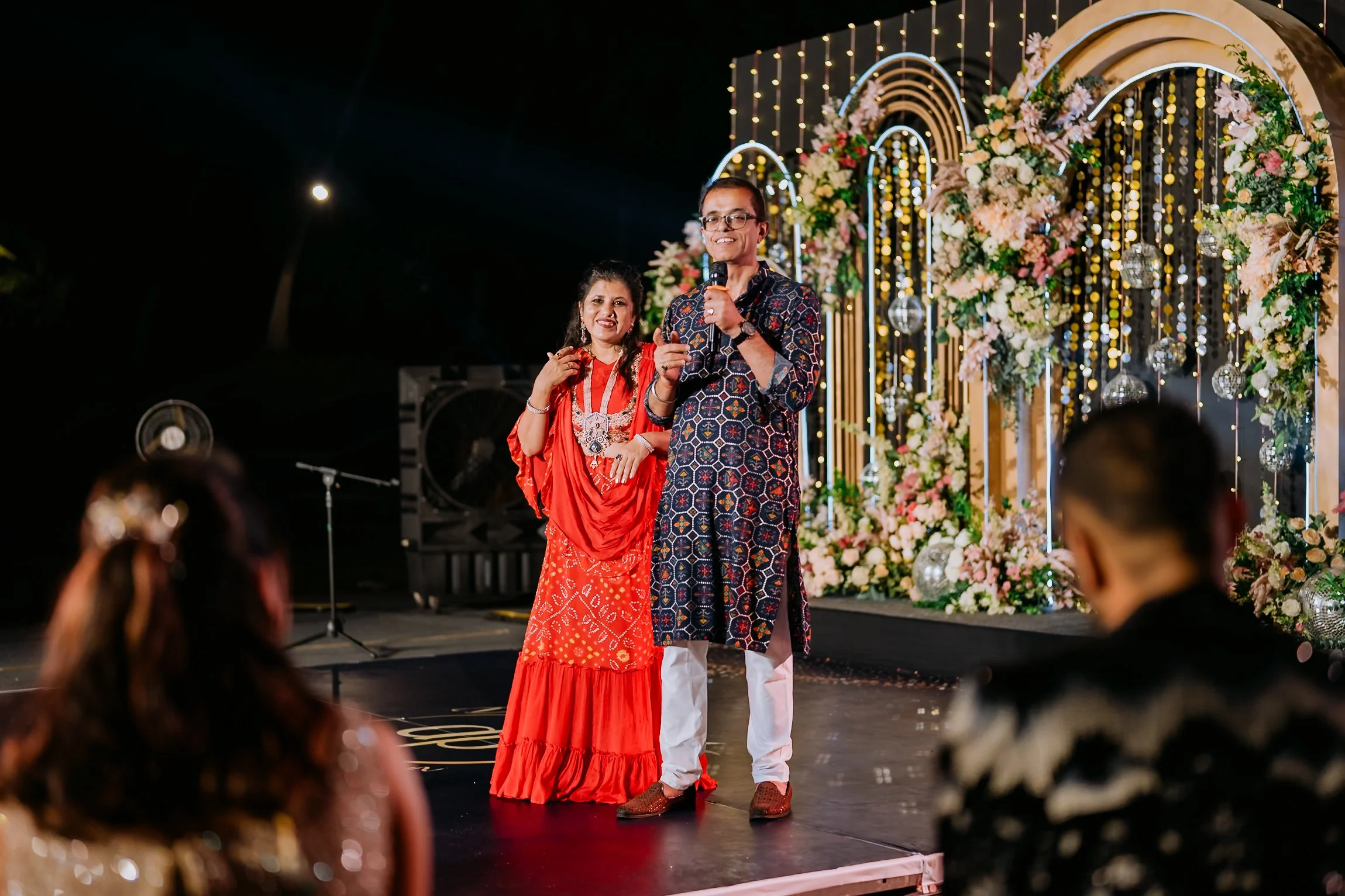 A man and a woman standing on a decorated outdoor stage at night, speaking into microphones during a celebration or wedding. The stage is adorned with flowers, greenery, and hanging lights.