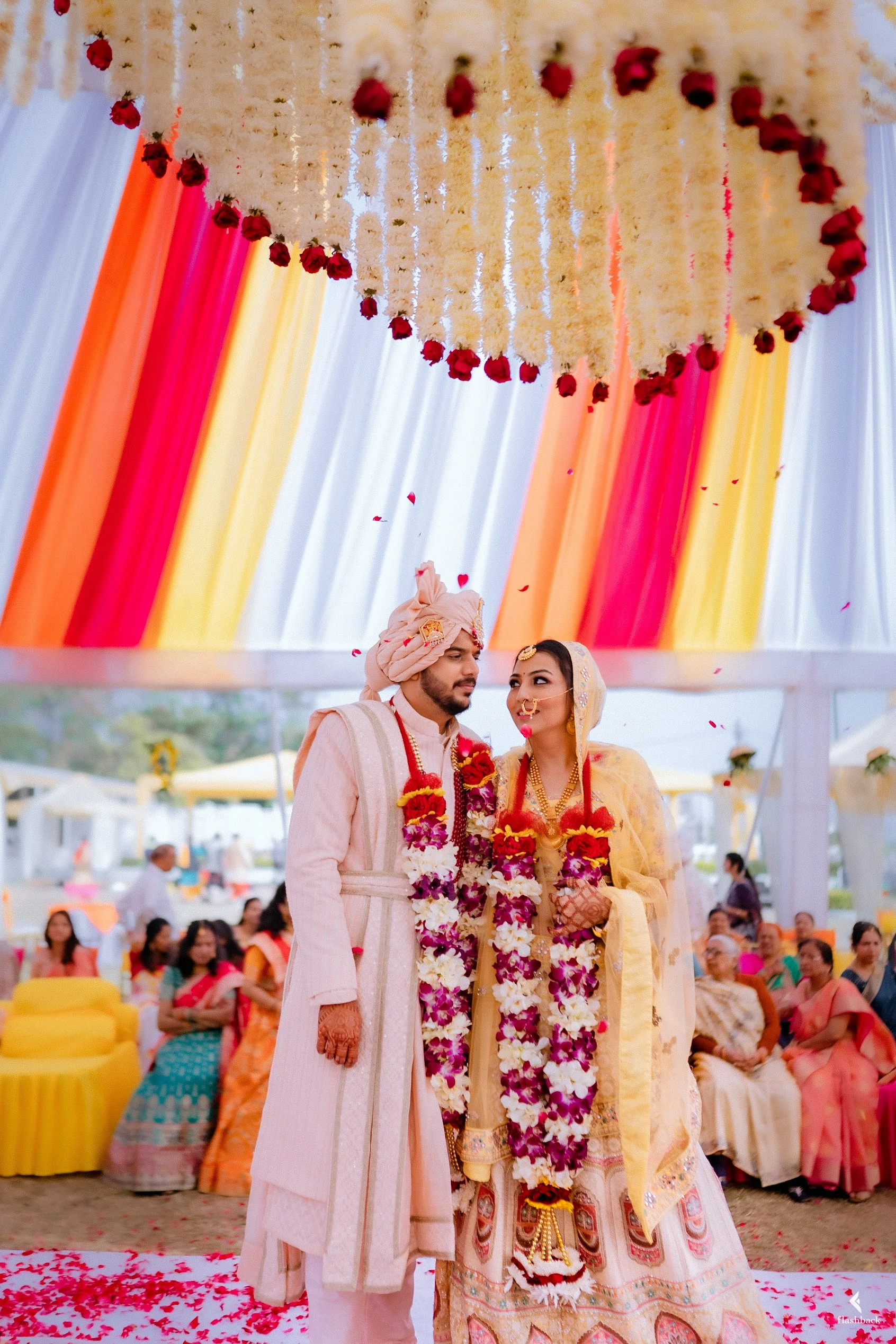 Indian bride and groom at wedding ceremony, decorated with flower garlands, under a floral canopy, guests seated around watching.