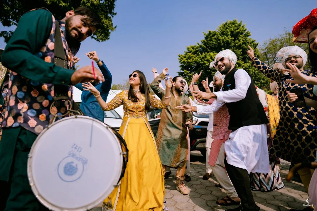 Group of people celebrating at an outdoor event, dancing and playing drums, dressed in traditional Indian attire, with trees and cars in the background.