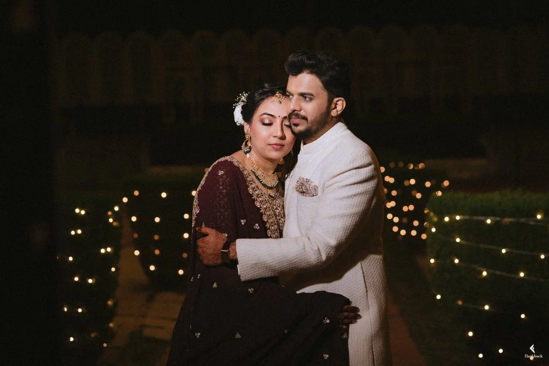 A groom in a white traditional Indian sherwani embraces a bride in an elegant maroon saree with gold embroidery, at a nighttime outdoor wedding with string lights in the background.