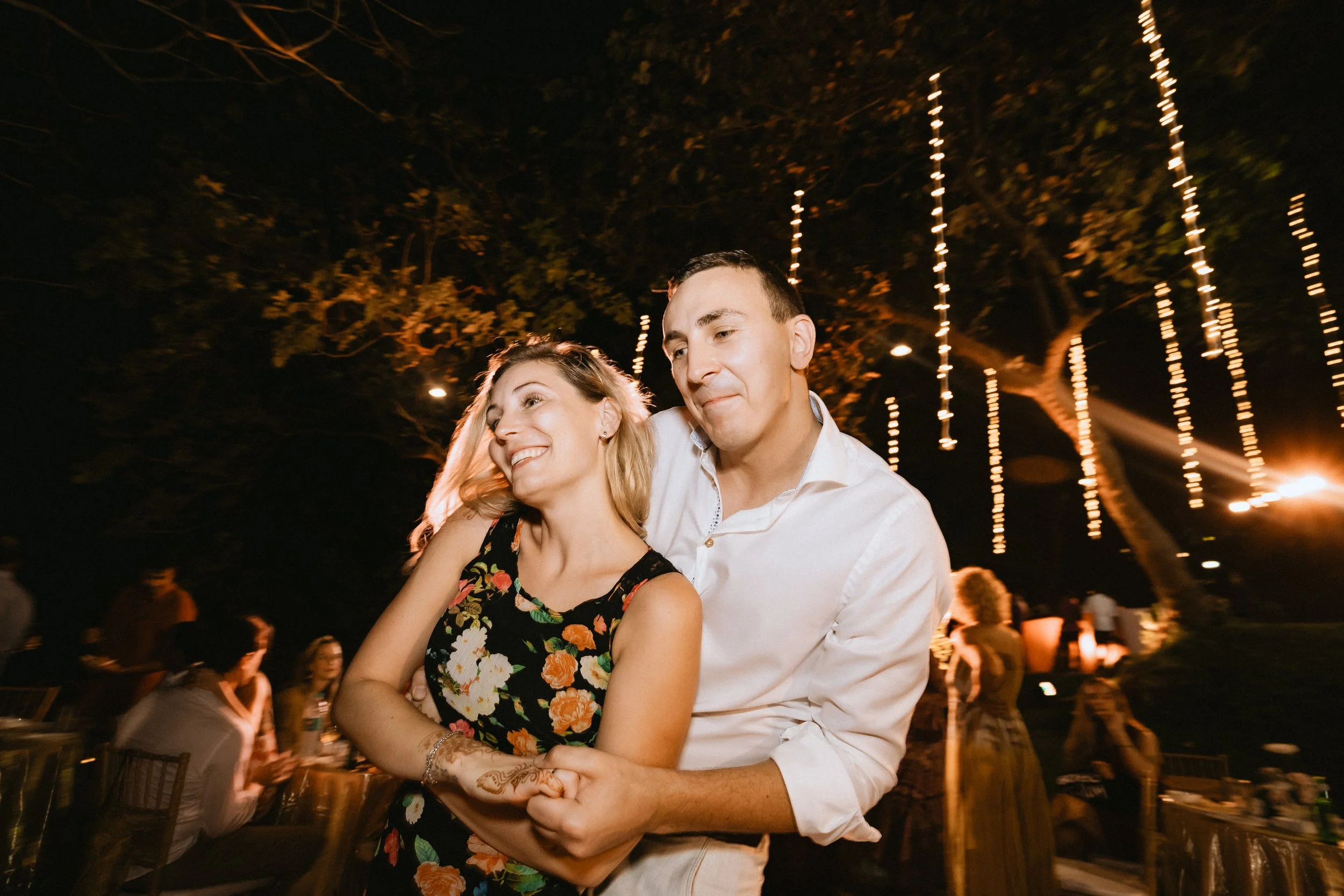 A smiling woman with curly hair and a black floral dress is being embraced by a man in a white shirt at a nighttime outdoor event with string lights and other guests in the background.